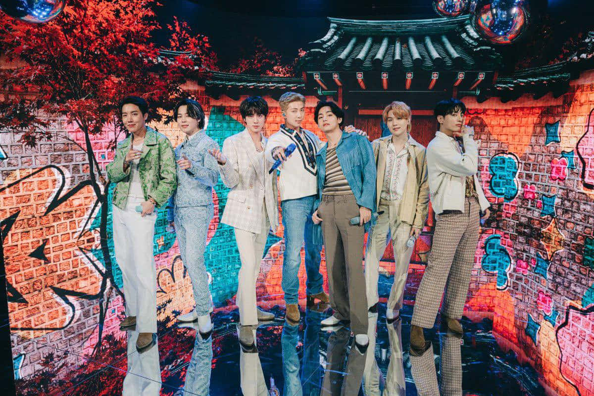 七位時尚K-pop表演者在色彩繽紛的塗鴉舞台上擺姿勢，背景有傳統屋頂。 — Bts 壁紙