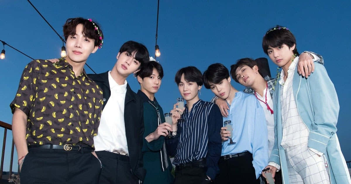 七位穿著時尚的年輕男子在黃昏時分於屋頂與串燈一起擺姿勢。 — Bts 壁紙