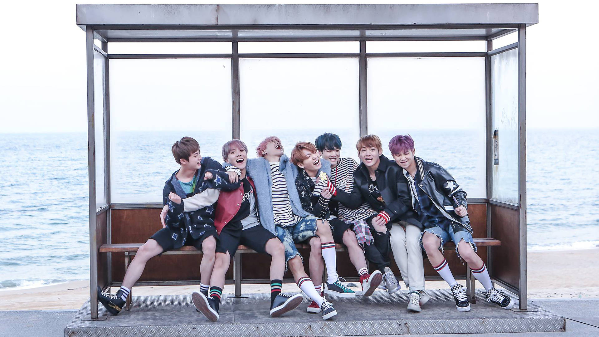 七位微笑的年輕男士穿著潮流服裝，坐在海邊巴士站長椅上。 — Bts 壁紙