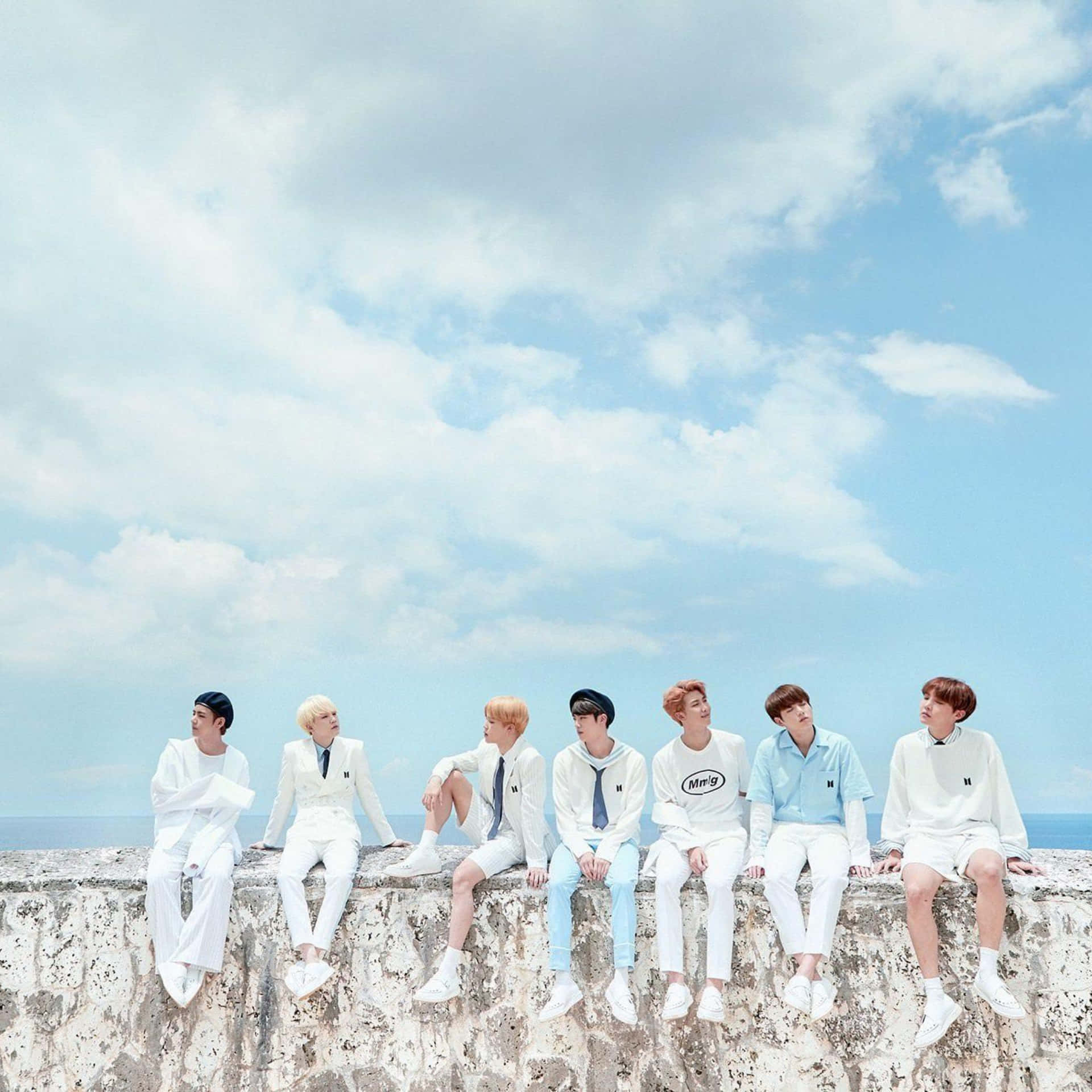  壁紙 — 七位身穿白色和淺藍色服裝的年輕男子，隨意地坐在風化的石牆上，背景是廣闊的藍天和蓬鬆的白雲。