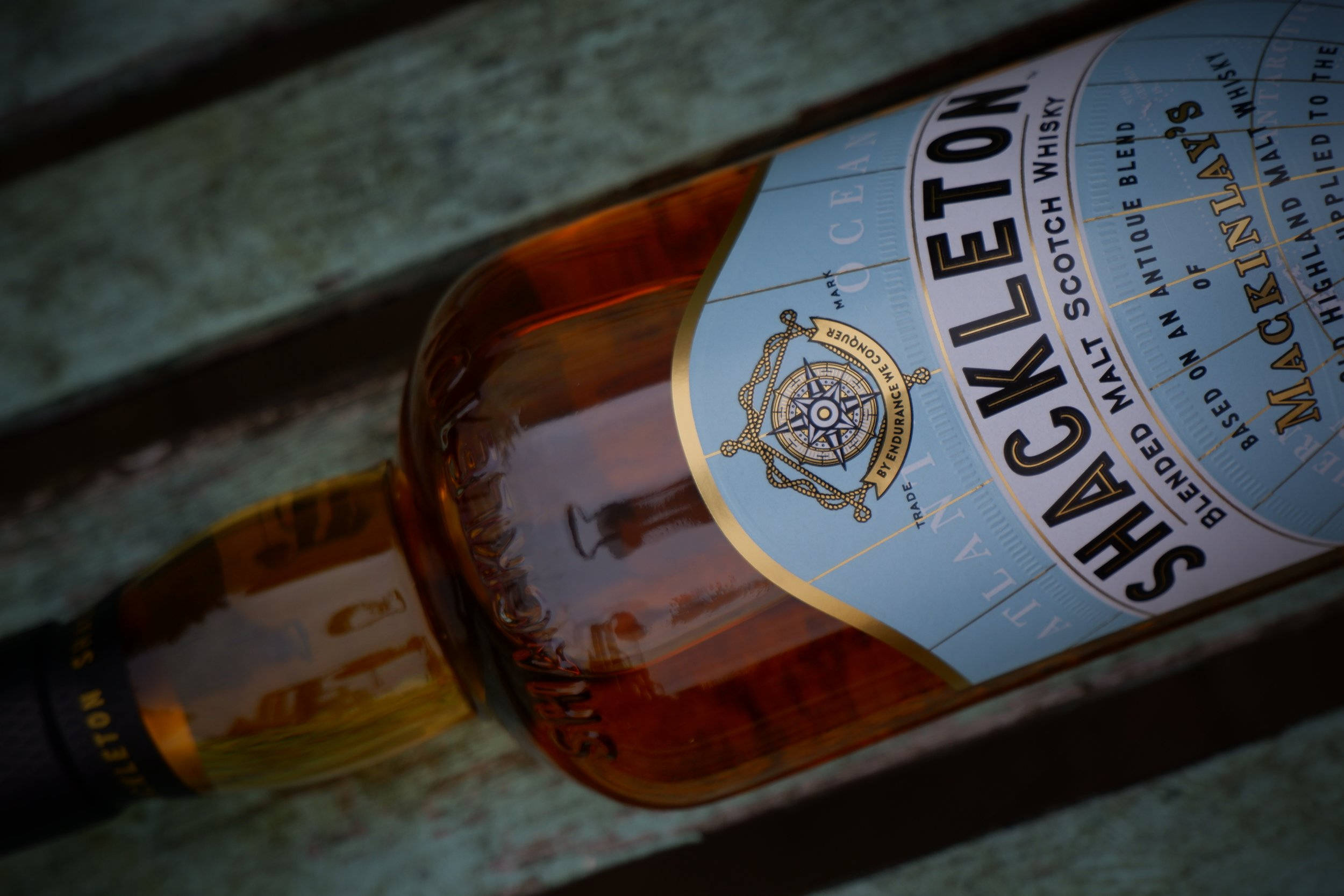 Drink 桌布 壁紙 — Shackleton 威士忌酒瓶的特寫，帶有淺藍色標籤和金色指南針標誌，放置在風化的木質表面上。