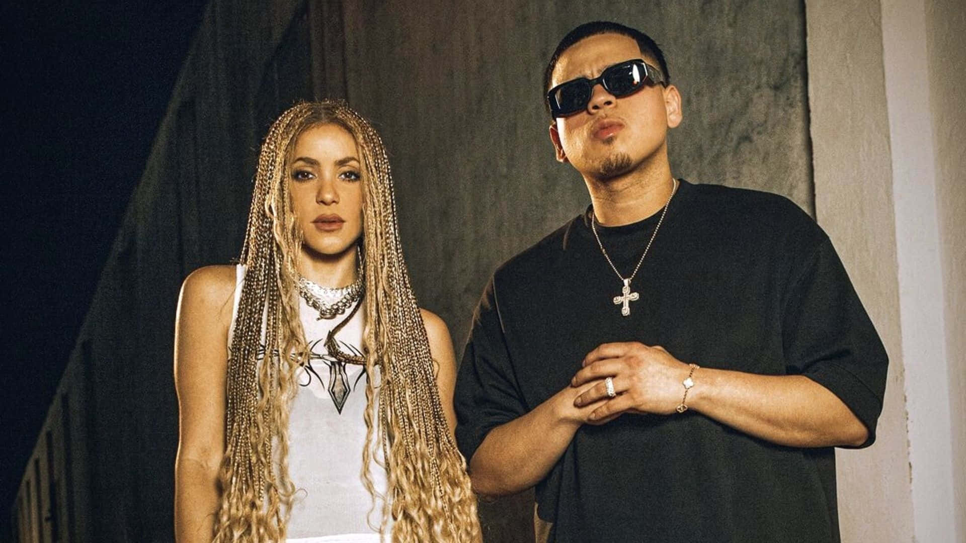  壁紙 — Shakira 和一個戴太陽眼鏡的男人在夜晚自信地靠在有紋理的混凝土牆上擺姿勢。