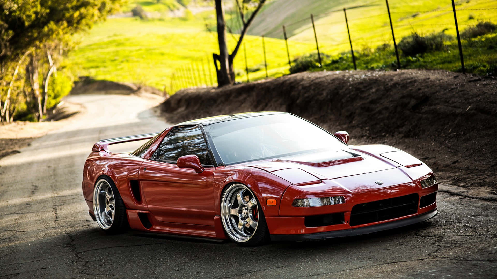 Honda Nsx 桌布 壁紙 — 一輛閃亮的紅色跑車配備客製化輪圈，停在一條蜿蜒的鄉村道路上，周圍是陽光明媚的綠色山丘和樹木。
