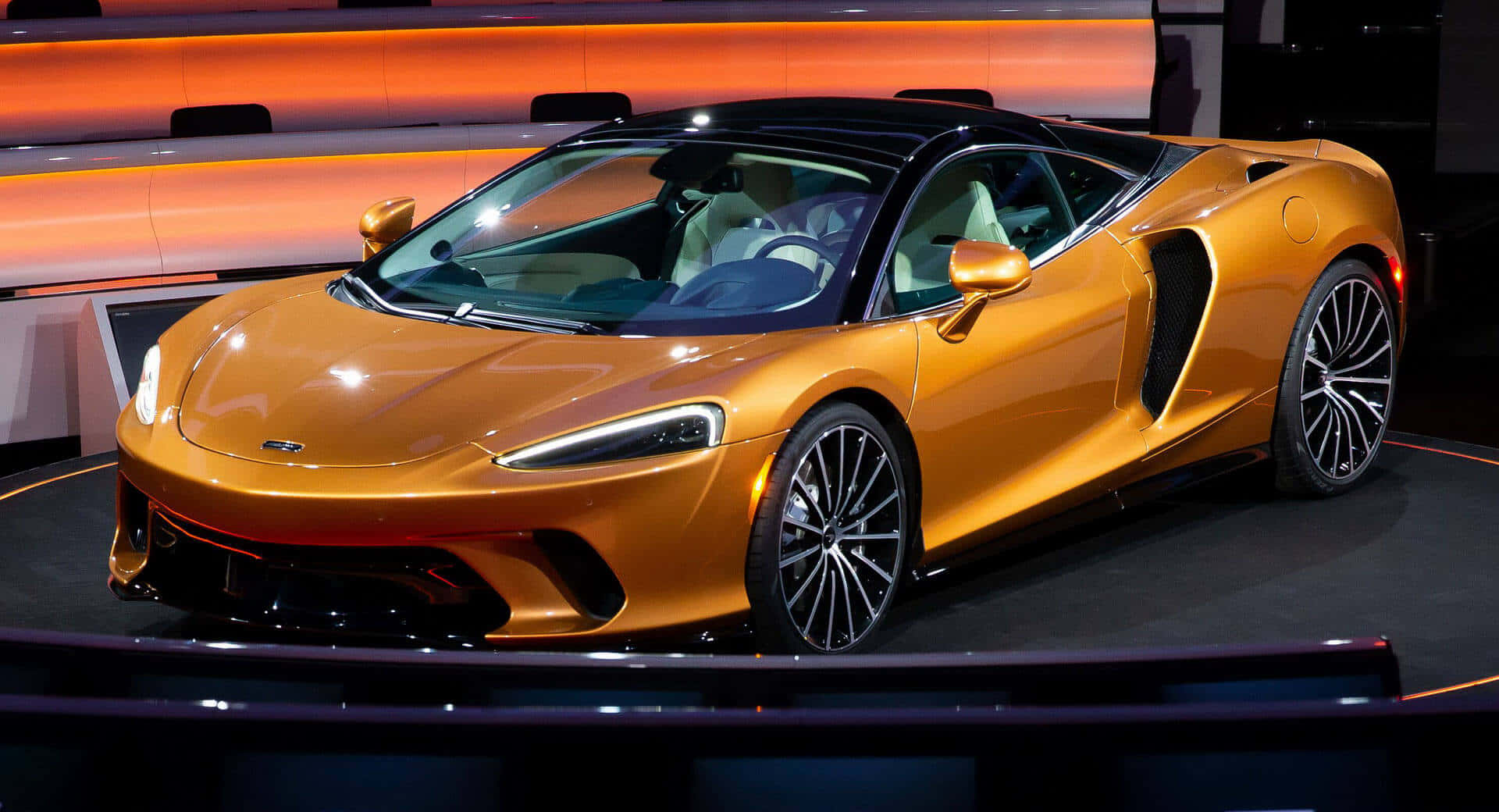 Mclaren Gt 桌布 壁紙 — 一輛閃亮的金色麥拉倫跑車，帶有黑色飾邊和時尚設計，在溫暖的橙色舞台燈光下展示。