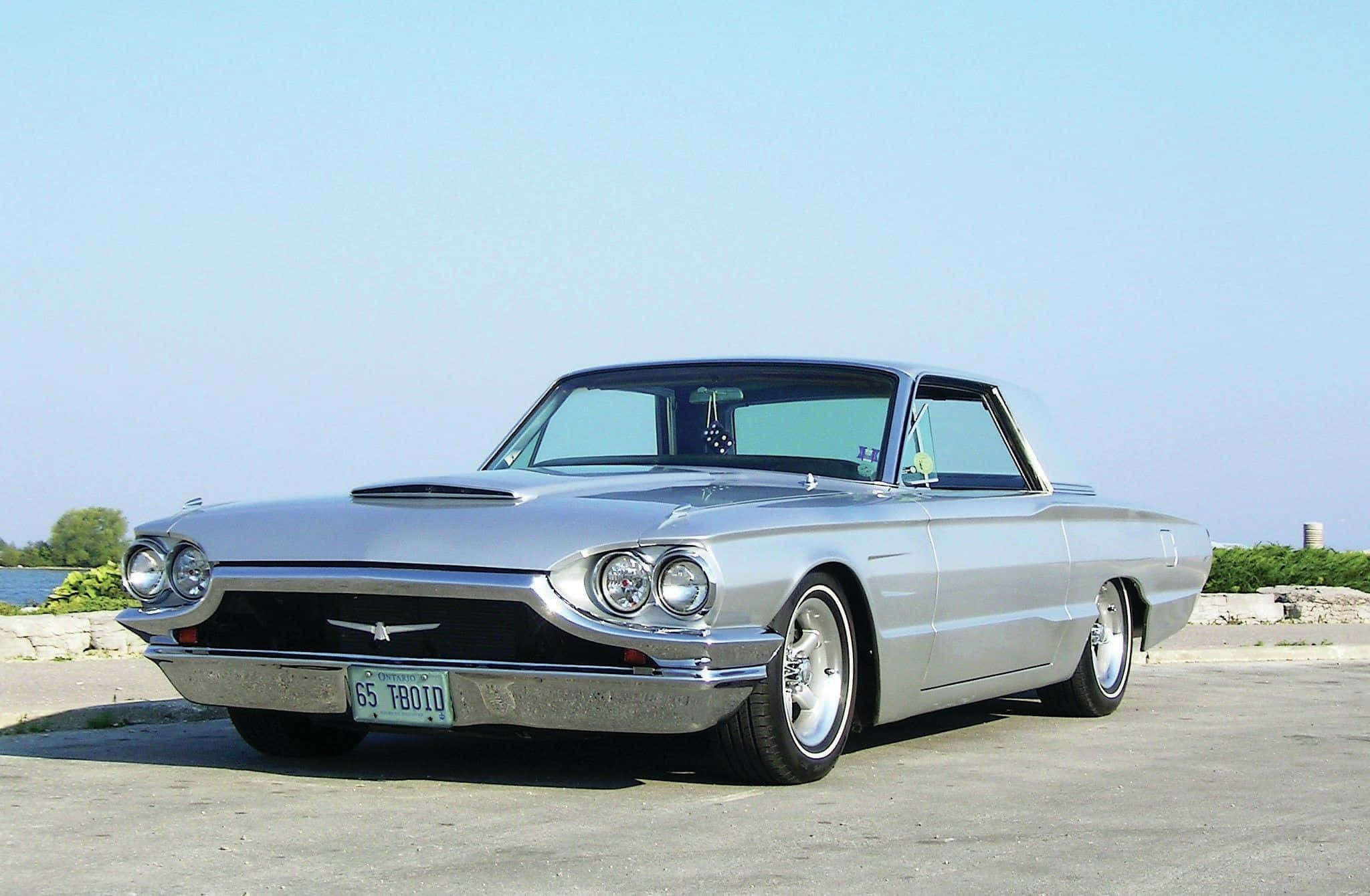 Ford Thunderbird 桌布 壁紙 — 一輛閃亮的銀色1965年福特雷鳥經典汽車停在柏油路上，背景是平靜的湖泊和綠植，上方是清澈的藍天。
