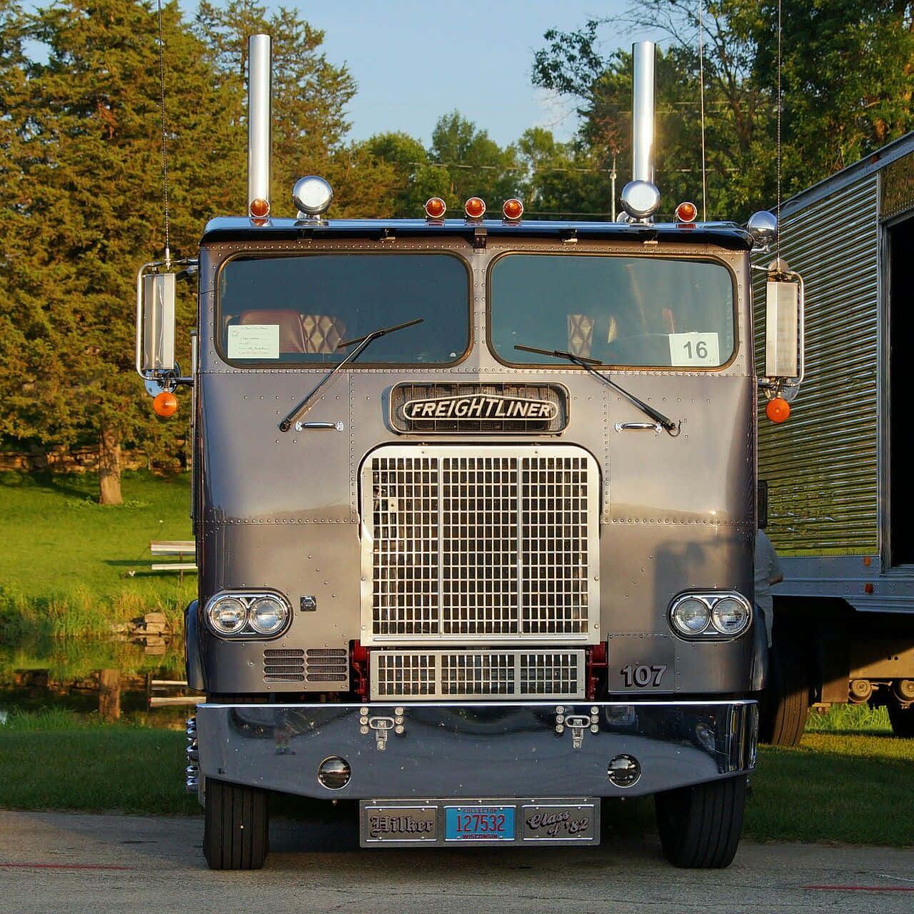 Kenworth T604 桌布 壁紙 — 一輛閃亮的銀色 Freightliner 半掛卡車，帶有鍍鉻裝飾和雙排氣管，停在鋪砌的停車場，旁邊是長滿草的池塘，正值黃金時刻。