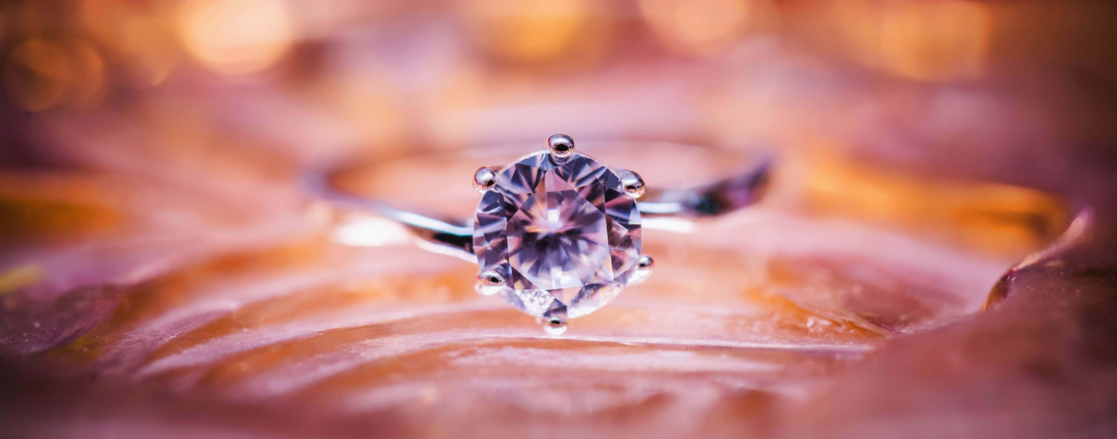 Purple Jewelry 桌布 壁紙 — 一枚閃爍的鑽石訂婚戒指在柔和的金色光線下，映襯著模糊的暖色調背景。