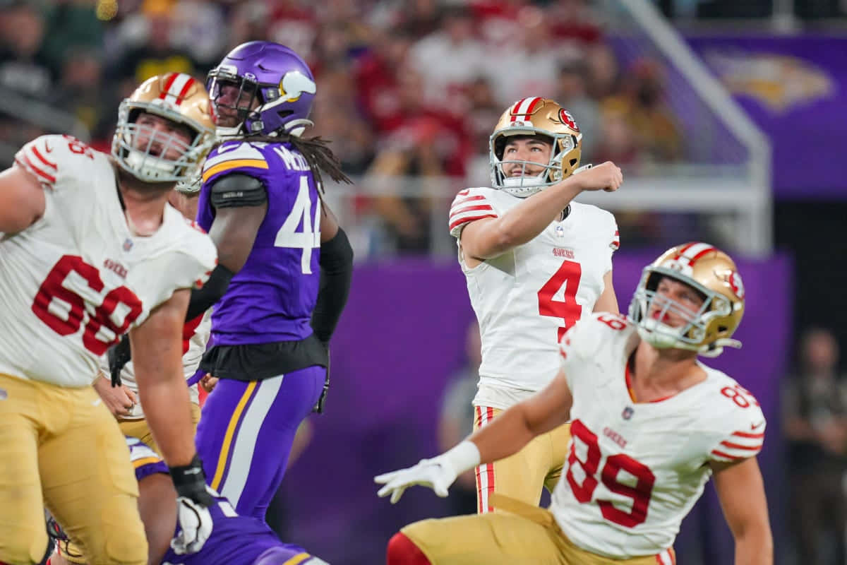  壁紙 — 身穿白色球衣的NFL四分衛傳球，紫色防守球員撲向，隊友在體育場內圍繞著他。