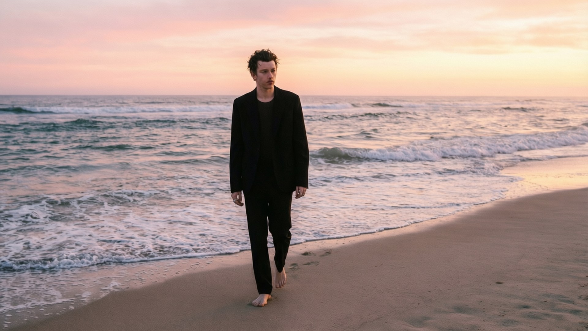 Sunrise Beach Walk 壁紙 — 一名身穿黑色西裝的男子赤腳走在沙灘上，溫柔的海浪在柔和的粉紅色和橙色日落天空下翻滾。