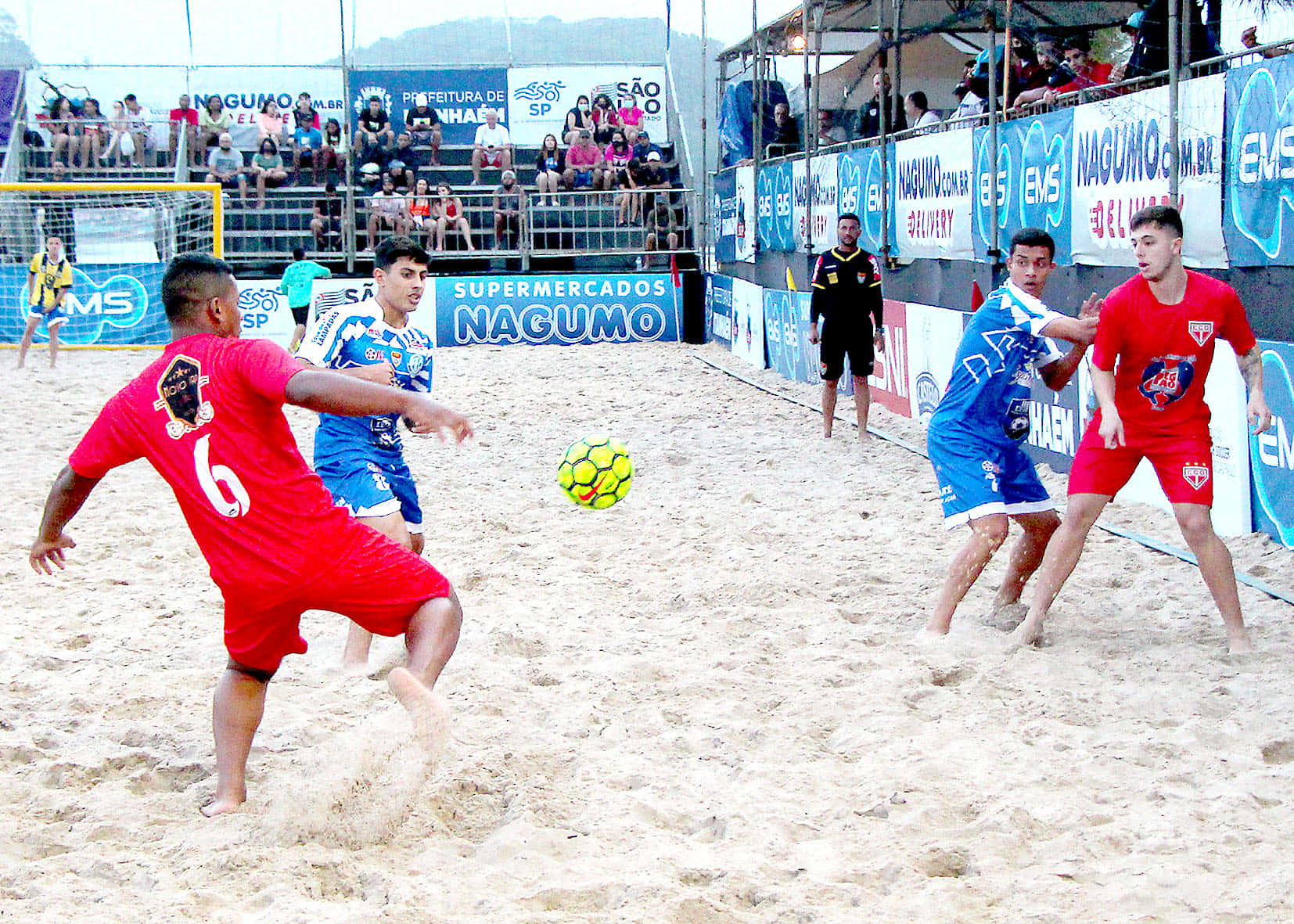 Beach Soccer 桌布 壁紙 — 身穿紅色和藍色球衣的球員在沙灘場地上激烈爭奪一個亮黃色的足球，觀眾在看台上觀看。