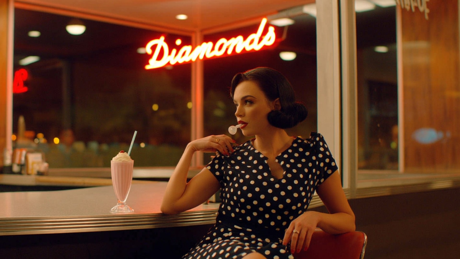 Vintage Diner Visit 壁紙 — 一位身穿黑白圓點連衣裙的女性在復古餐廳裡，在寫著Diamonds的發光霓虹燈下喝著奶昔。