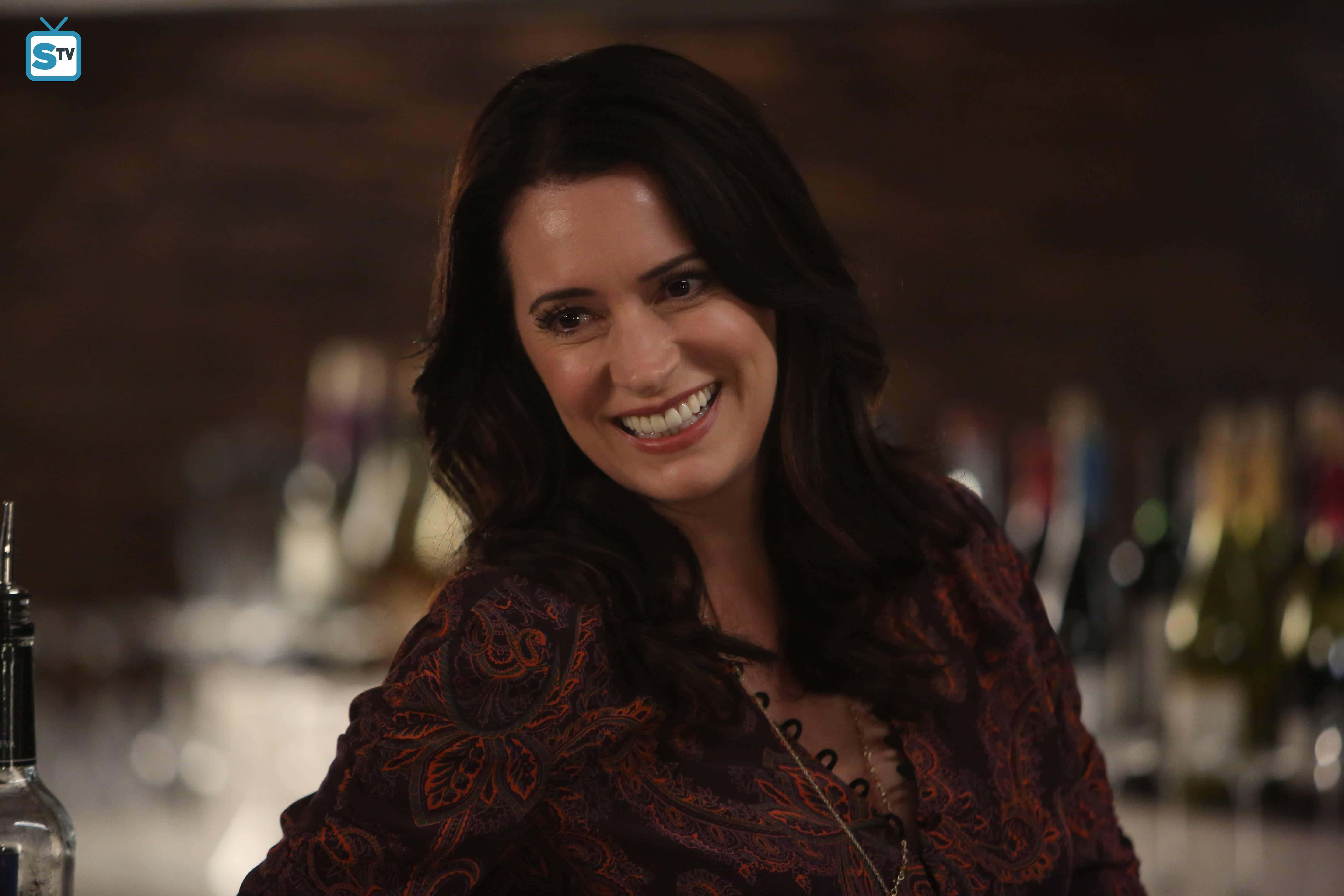 paget brewster 桌布 壁紙 — 一位深色頭髮的女性在燈光昏暗的酒吧中，背景有模糊的酒瓶，對著鏡頭溫暖微笑。