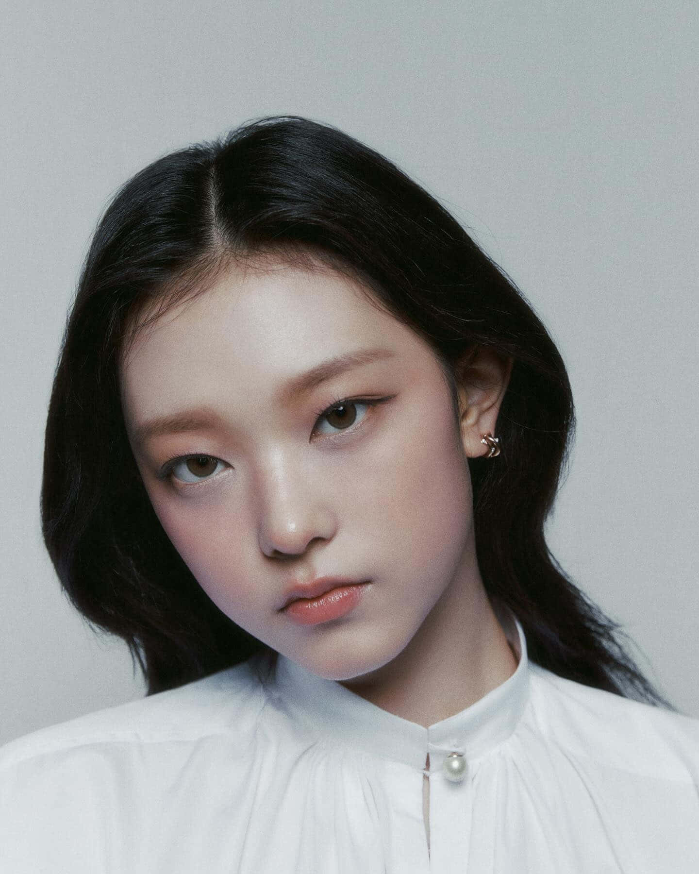  壁紙 — 一位深色頭髮、妝容淡雅的年輕女性，穿著白色珍珠鈕扣領襯衫，在純灰色背景前直視鏡頭。