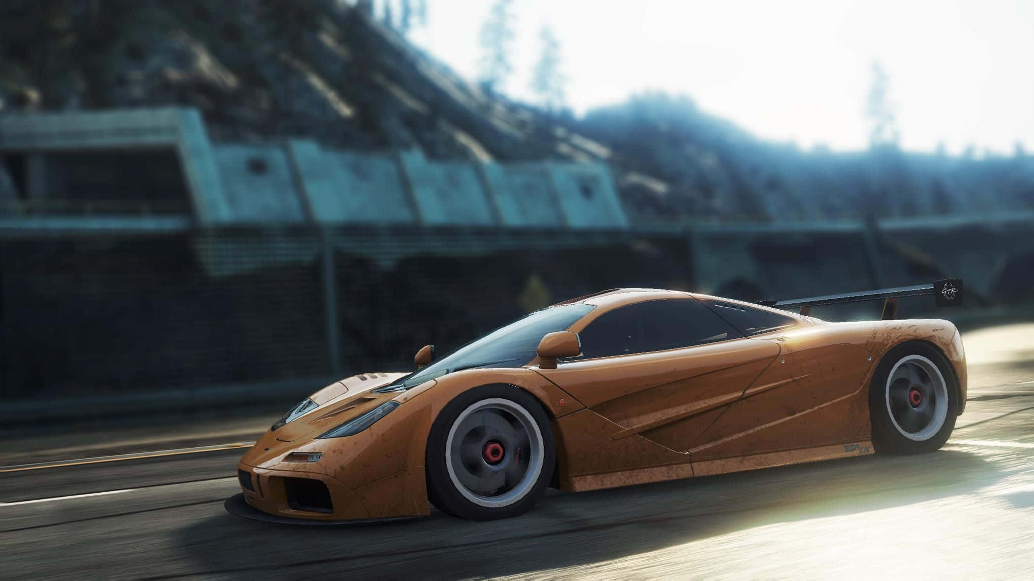 Mclaren F1 桌布 壁紙 — 一輛生鏽的橙色McLaren F1賽車在陽光明媚的賽道上疾馳，背景有模糊的動感和混凝土護欄。