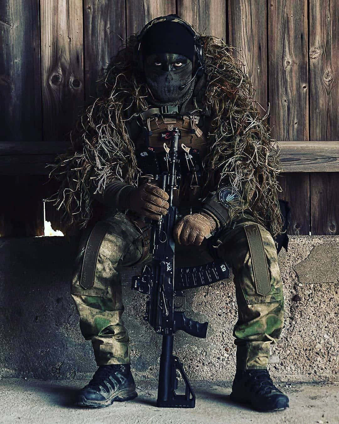  壁紙 — 一名身穿全套迷彩服和吉利服的士兵蹲在風化的木牆旁，手持突擊步槍。