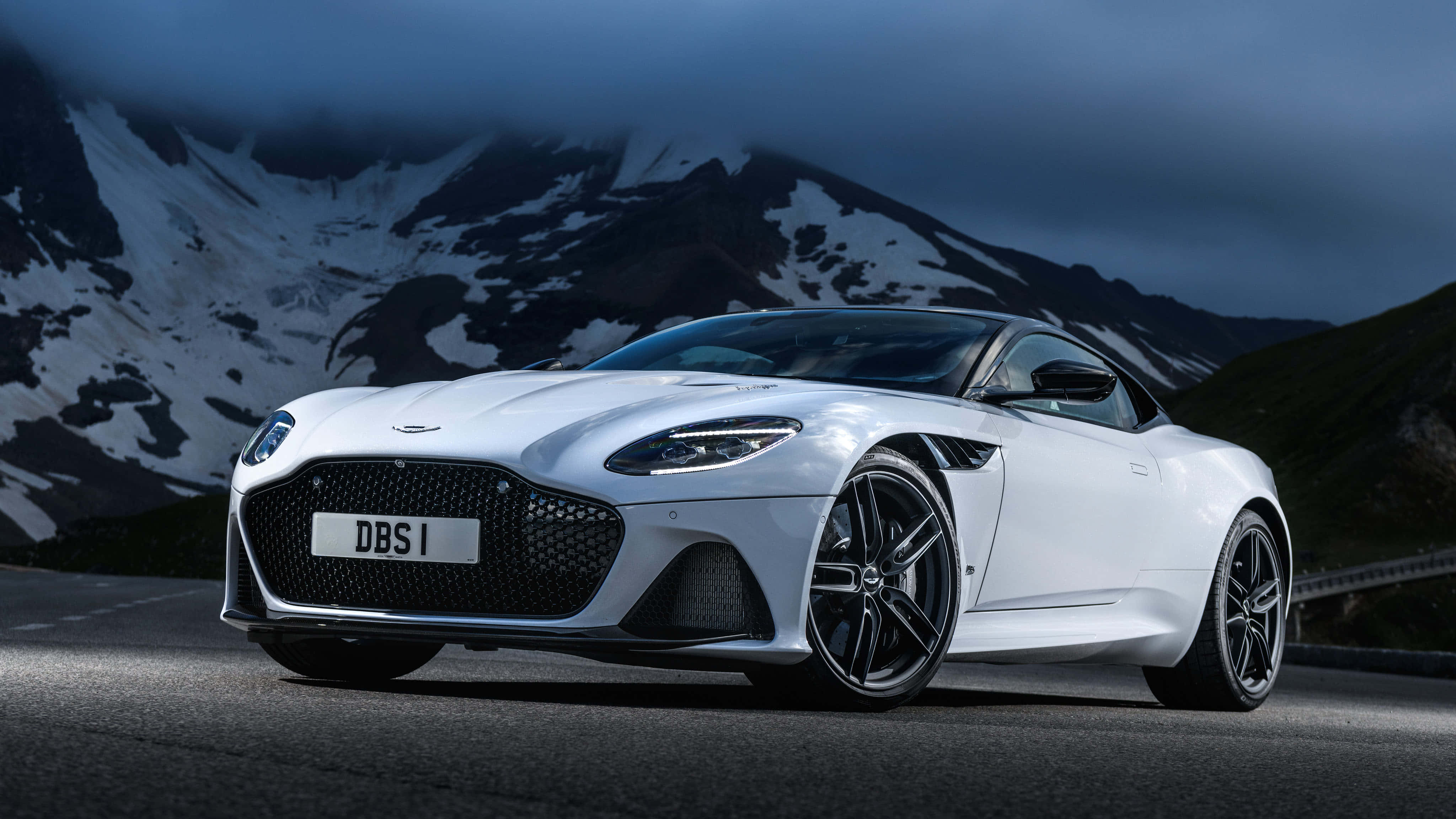 Aston Martin Dbs Superleggera 桌布 壁紙 — 一輛時尚的白色Aston Martin DBS跑車停在山路上，背景是白雪覆蓋的山峰，天空呈現陰沉的暮色。