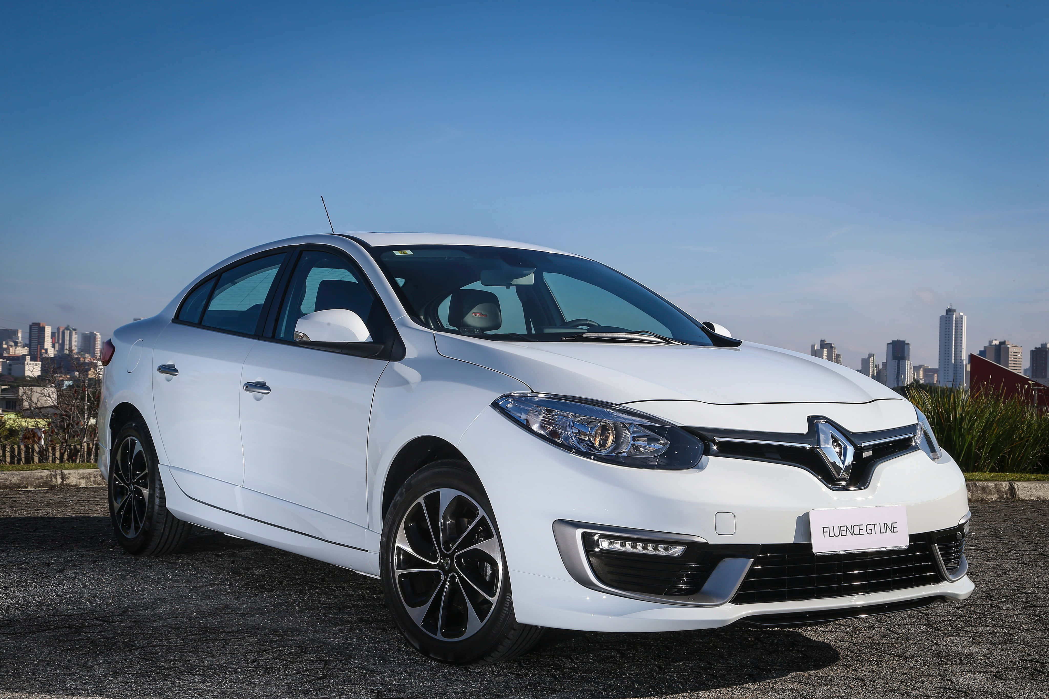 Renault Fluence 桌布 壁紙 — 一輛時尚的白色雷諾 Fluence GT Line 轎車停在碎石路上，背景是晴朗藍天下的城市天際線。