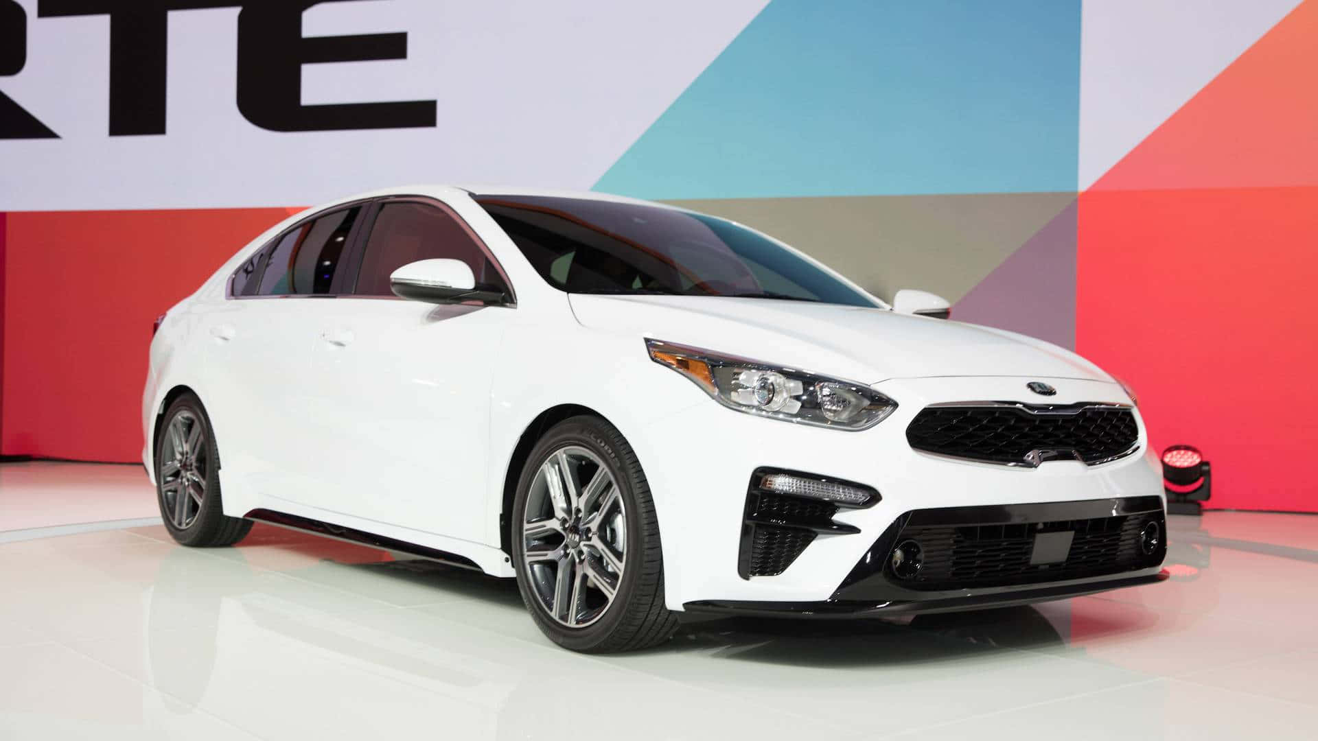 Kia Forte 桌布 壁紙 — 一輛時尚的白色起亞轎車在明亮的燈光下，在光亮的展廳地板上閃閃發光，背景是色彩繽紛的幾何圖案。