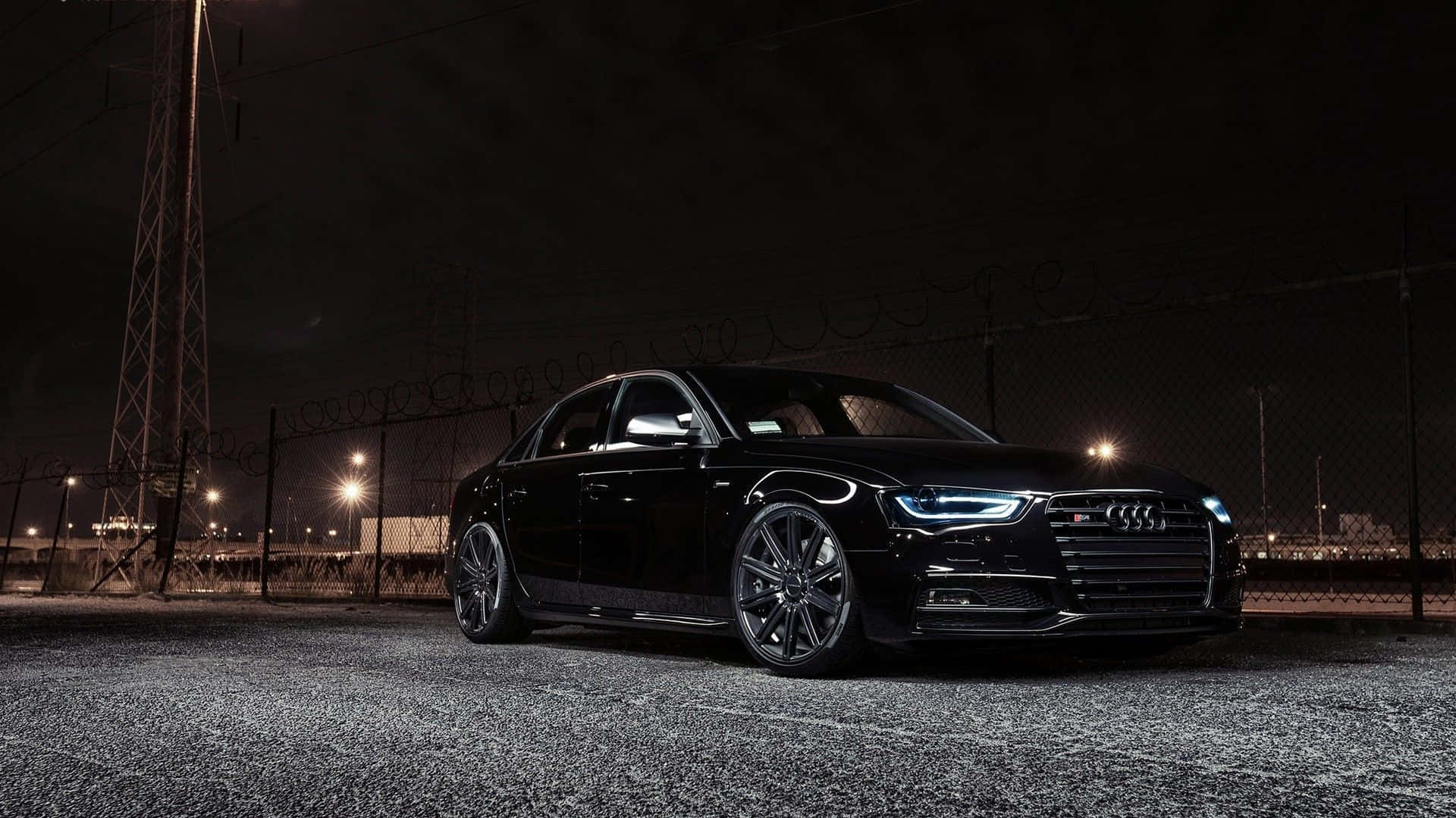 Audi A4 桌布 壁紙 — 一輛時尚的黑色奧迪轎車在夜燈下閃閃發光，停在帶有鐵絲網的鏈條圍欄旁。