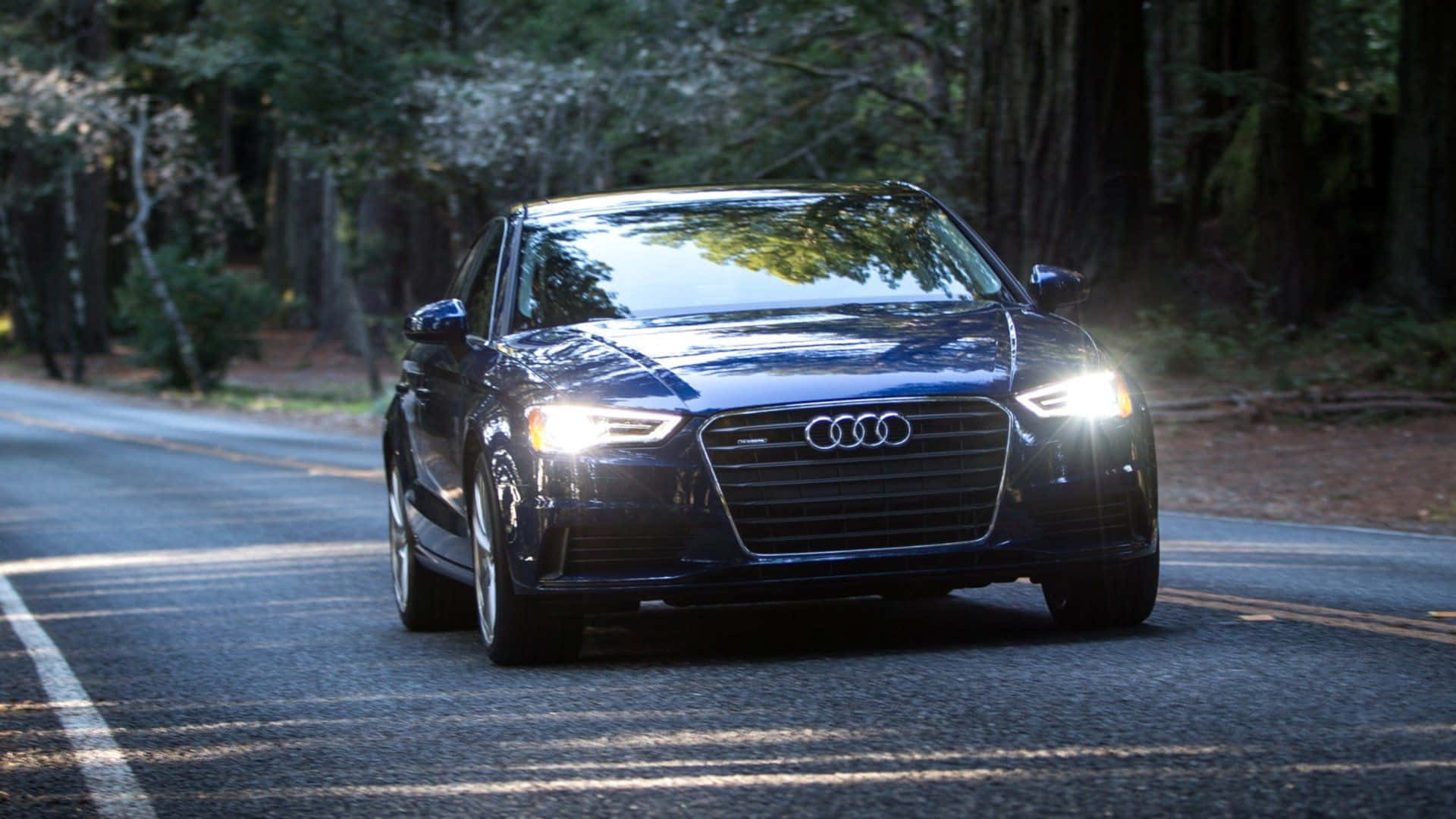 Audi A3 桌布 壁紙 — 一輛時尚的黑色奧迪轎車在樹林環繞的道路上駛向鏡頭，車頭燈在斑駁的陽光中閃耀。