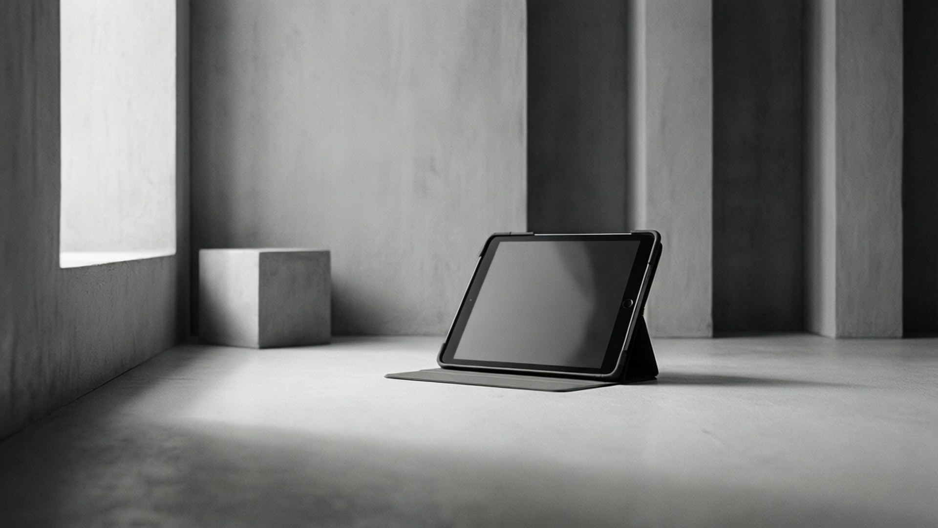 Minimalist Tablet 壁紙 — 一部時尚的黑色平板電腦放在支架上，位於簡約的混凝土房間內，窗戶透進柔和自然光。