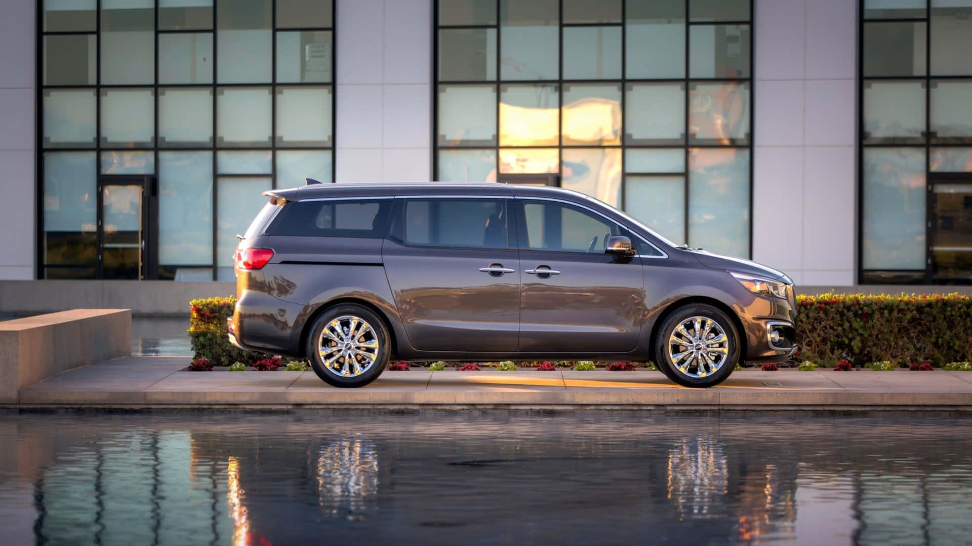 Kia Sedona 桌布 壁紙 — 一輛時尚的灰色小型貨車停放在現代玻璃建築前的反射水景旁邊。