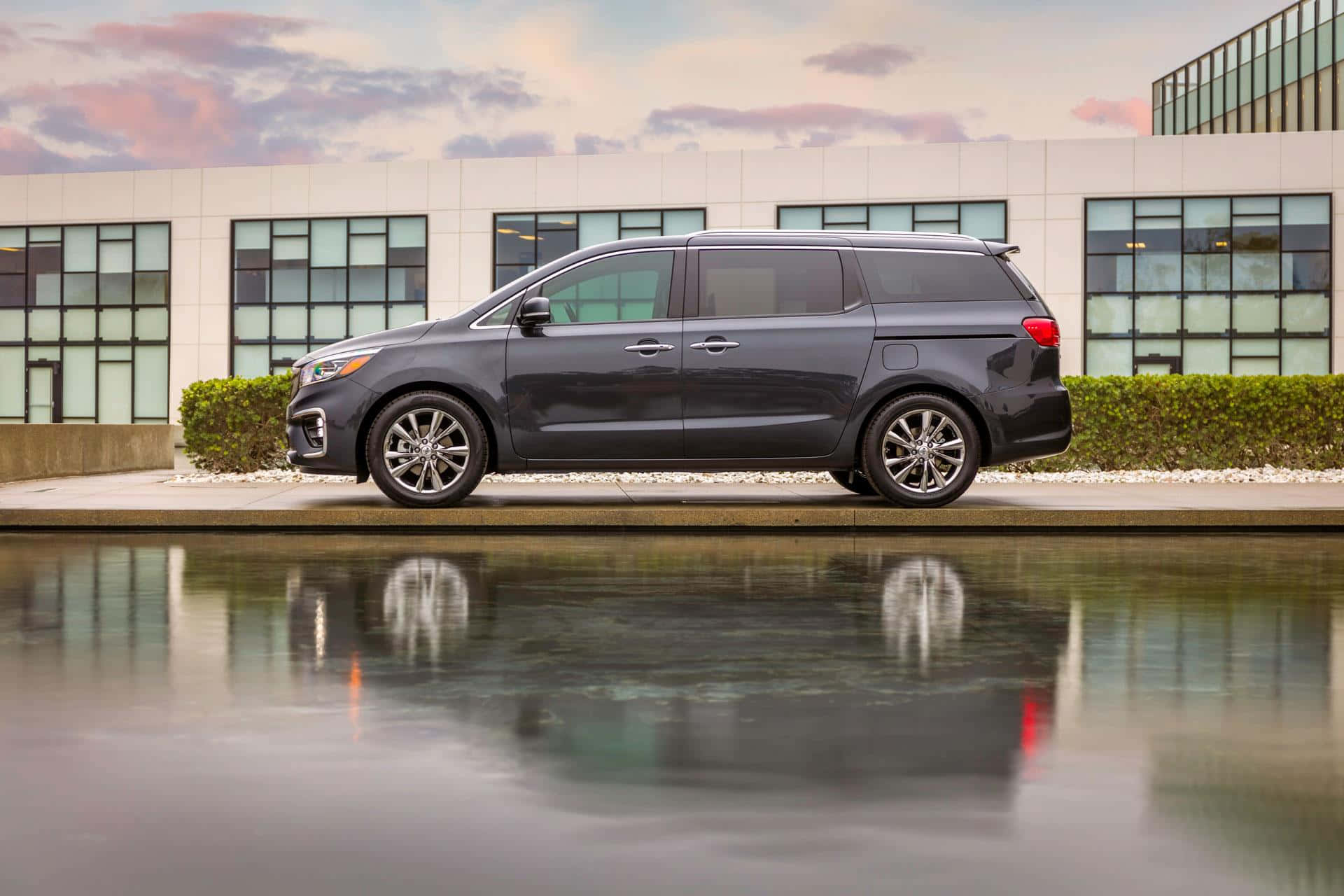 Kia Sedona 桌布 壁紙 — 一輛時尚的深灰色小型貨車停泊在黃昏時分現代玻璃建築前的反射水景旁邊。