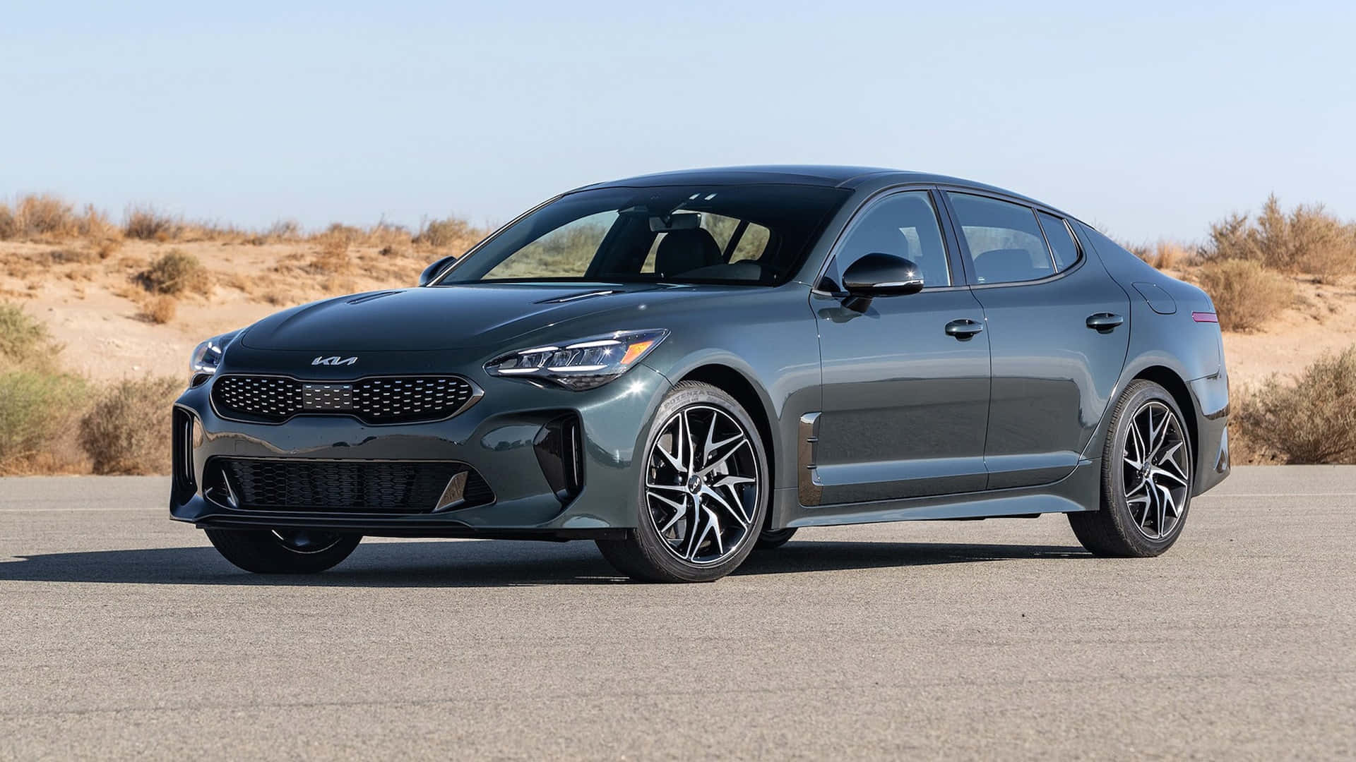 Kia Stinger 桌布 壁紙 — 一輛時尚的啞光灰色Kia Stinger運動轎車停在瀝青路上，背景是沙漠灌木和晴朗的藍天。