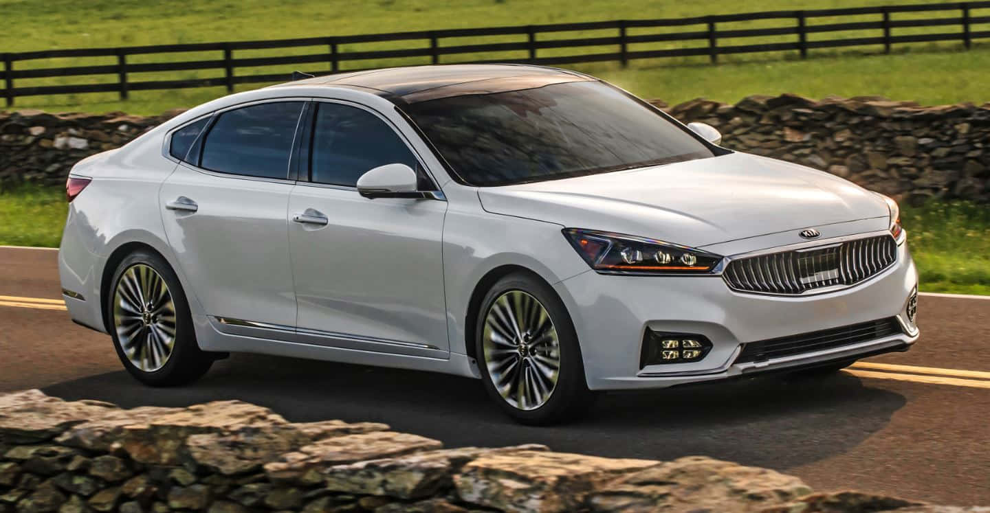Kia Cadenza 桌布 壁紙 — 一輛時尚的銀色轎車沿著蜿蜒的鄉村道路行駛，道路兩旁是石牆和翠綠的田野。