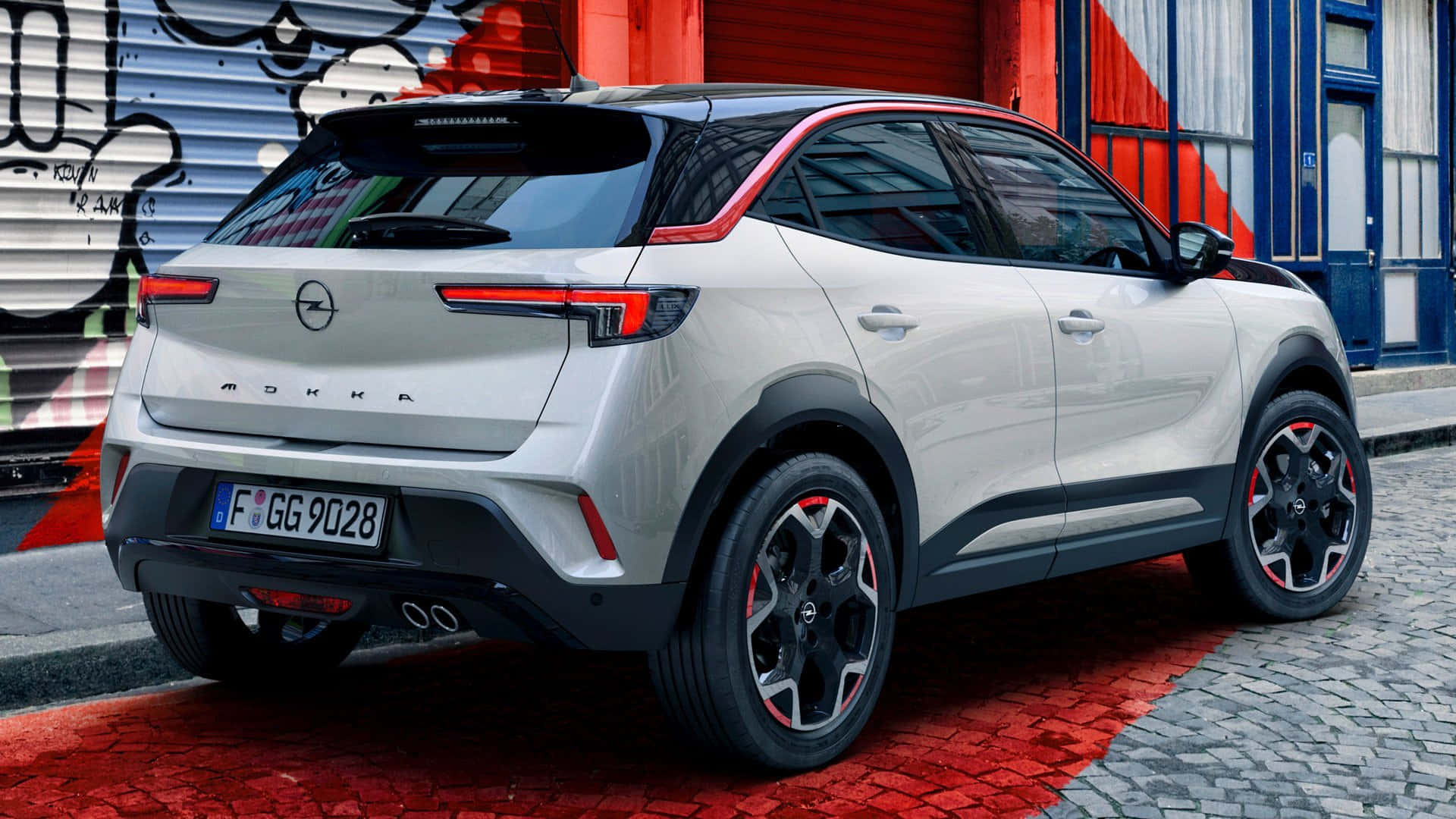 Opel Mokka 桌布 壁紙 — 一輛時尚的銀色Opel Mokka SUV停放在紅色鵝卵石街道上，旁邊是佈滿塗鴉的工業建築。