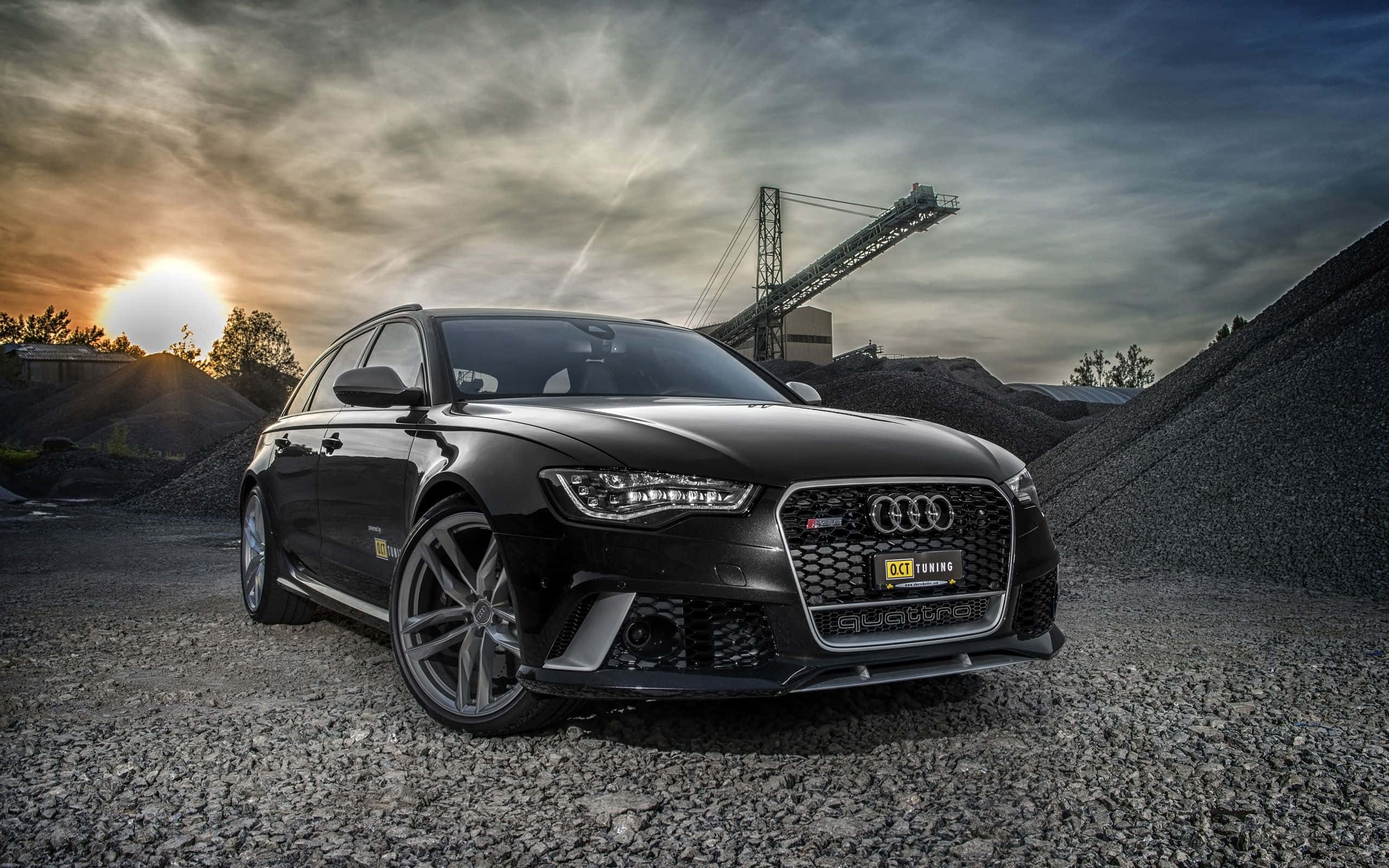 Audi S6 桌布 壁紙 — 一架時尚黑色Audi RS6 Avant停放在採石場的礫石上，背景有壯麗的日落和工業輸送帶結構。
