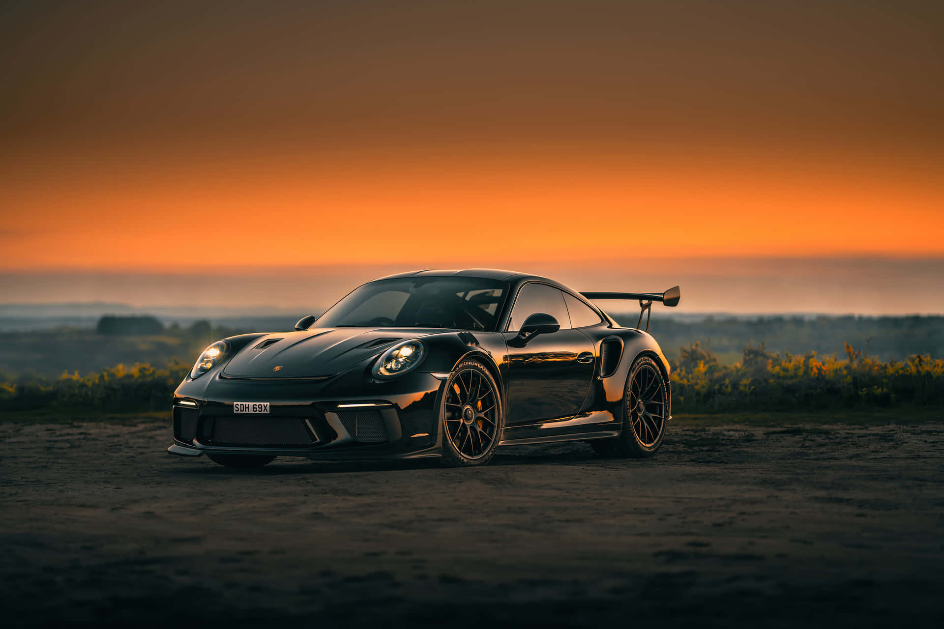  壁紙 — 一輛時尚黑色保時捷911 GT2 RS停在日落時分的泥土路上，天空呈現火熱橙色，遠方有山丘。