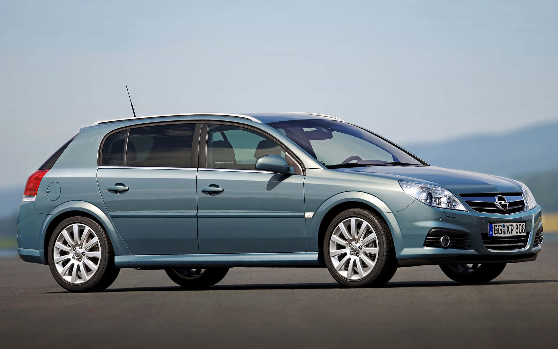 Opel Signum 桌布 壁紙 — 一輛時尚的淺藍色歐寶雅特掀背車停在開闊的瀝青停車場上，背景是晴朗天空下的朦朧山脈。