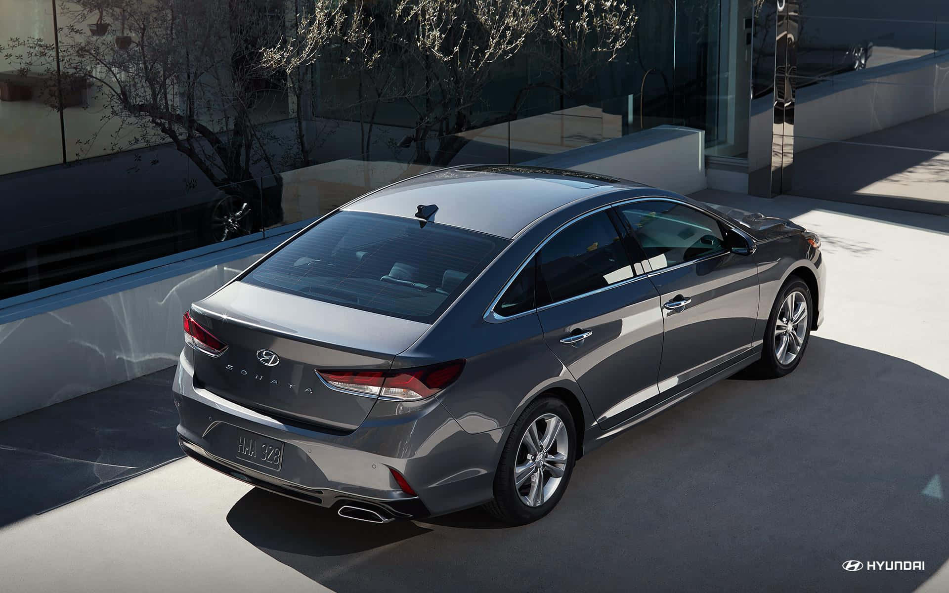 Hyundai Sonata 桌布 壁紙 — 一架時尚的銀色現代索納塔轎車停放在現代混凝土車道上，背景有玻璃牆和光禿禿的樹木。