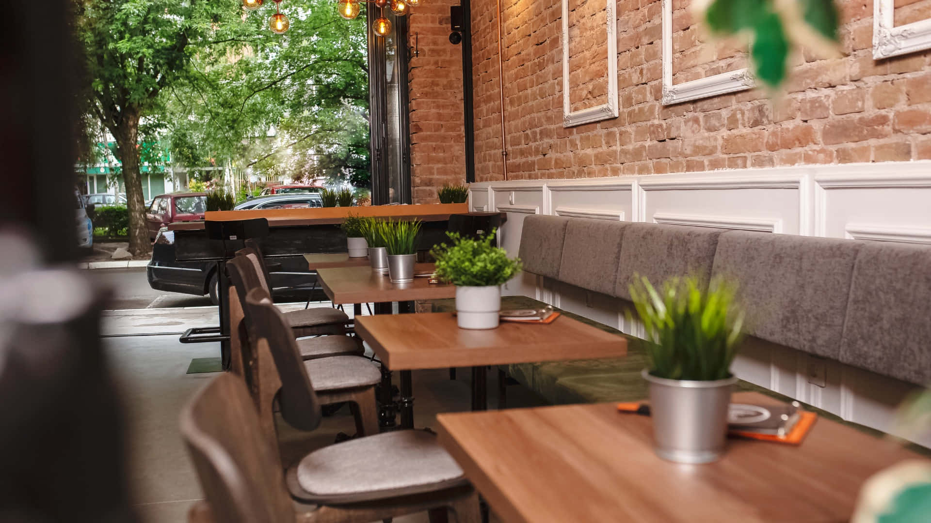 Coffee Shop 桌布 壁紙 — 舒適咖啡館內部，設有木桌、灰色卡座、裸露磚牆和綠色盆栽植物，在溫暖的吊燈下。