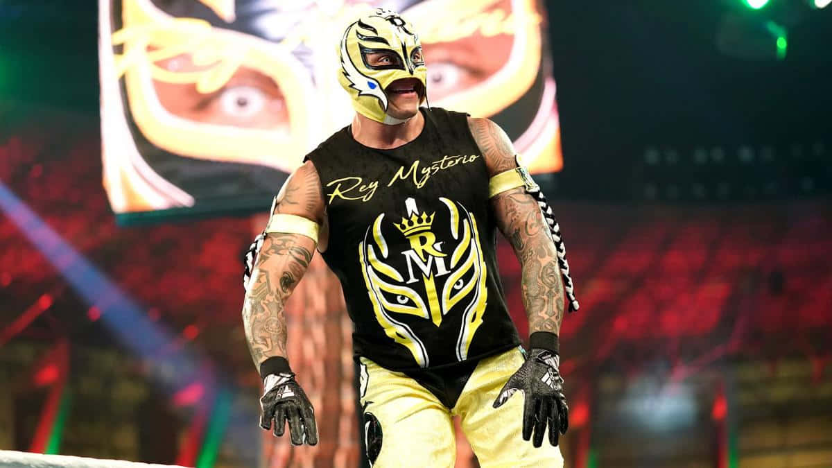  壁紙 — 摔角手 Rey Mysterio 戴著標誌性的黃黑 lucha libre 面具和裝備，在明亮的場館燈光下站在擂台上。