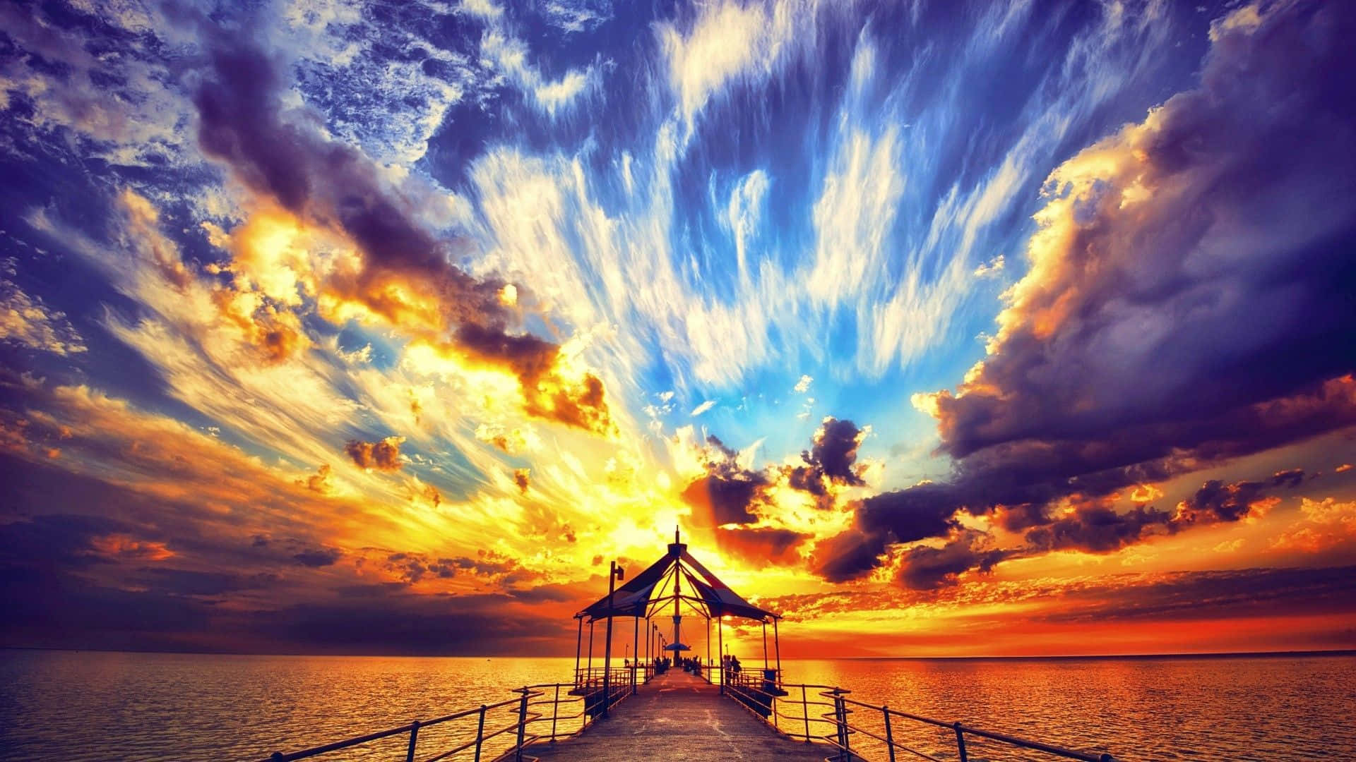 Colorful Sunset 桌布 壁紙 — 水上戲劇性的日落，碼頭通向涼亭，天空有火橙色和深藍色的雲彩。