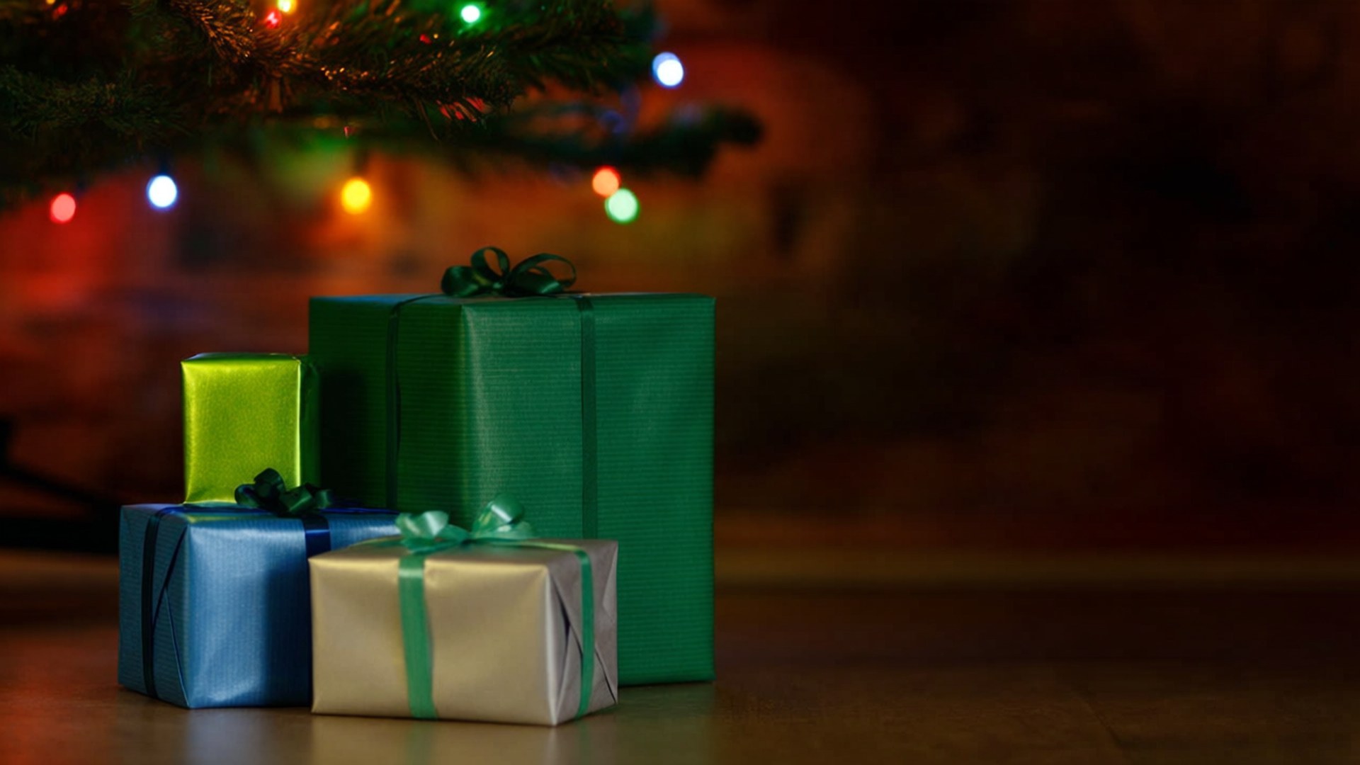 Gifts Under Tree 壁紙 — 四個節日包裝的禮物，綠色、藍色和銀色，放在發光的聖誕樹枝下。