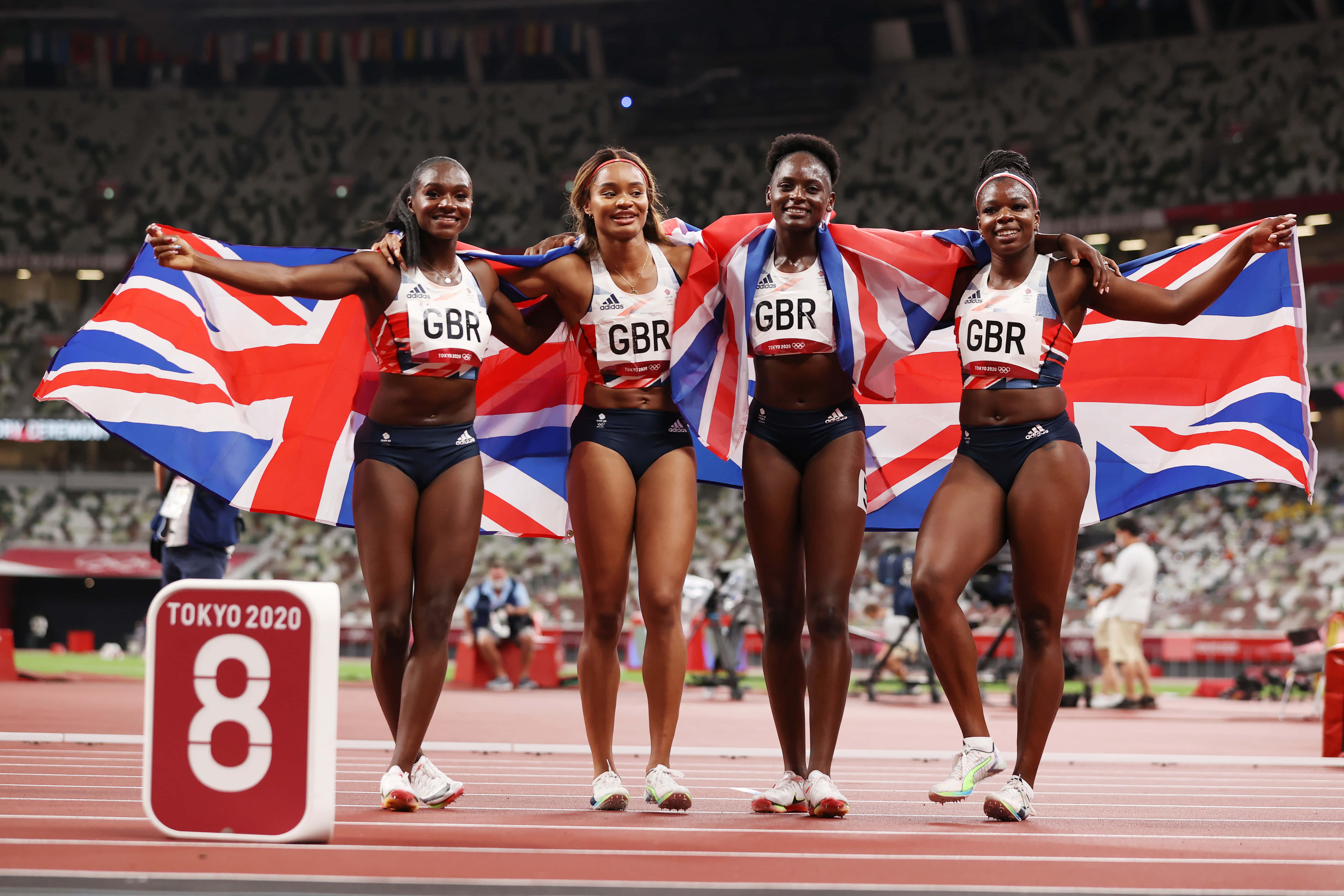 sport 桌布 壁紙 — 四位英國女運動員在跑道上慶祝，手持一面大的英國國旗，附近有東京2020標誌。