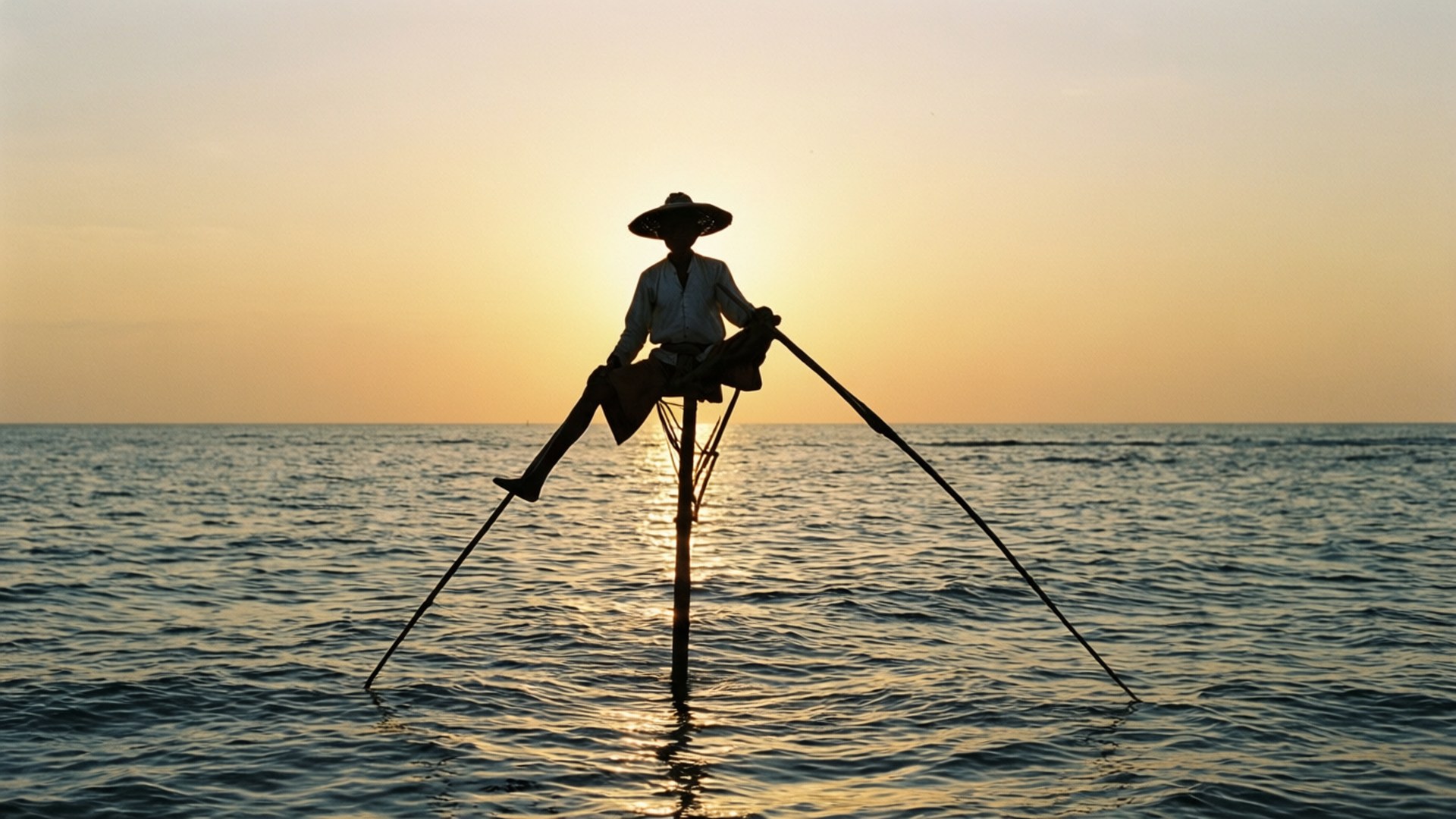 Stilt Fisherman at Sunset 壁紙 — 漁夫在金色日落時平靜的海水中平衡在桿上的剪影。