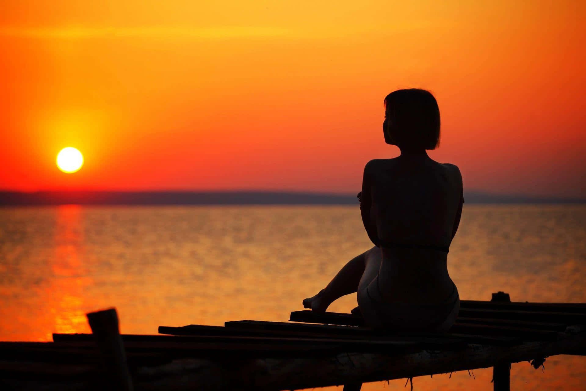 Sunset Silhouette 桌布 壁紙 — 一個女人獨自坐在木碼頭上，看著夕陽在平靜的水面上落下。