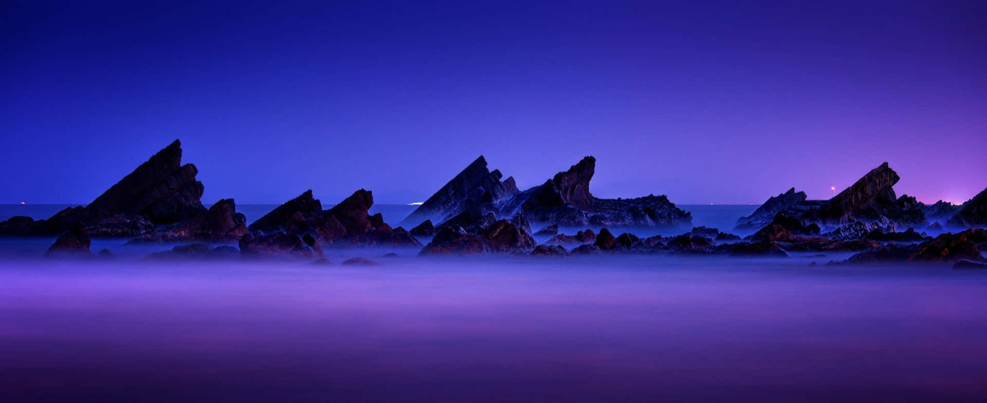 鋸齒狀岩石從霧氣瀰漫的紫色海洋中在暮色天空下浮現 — 3440X1440 壁紙