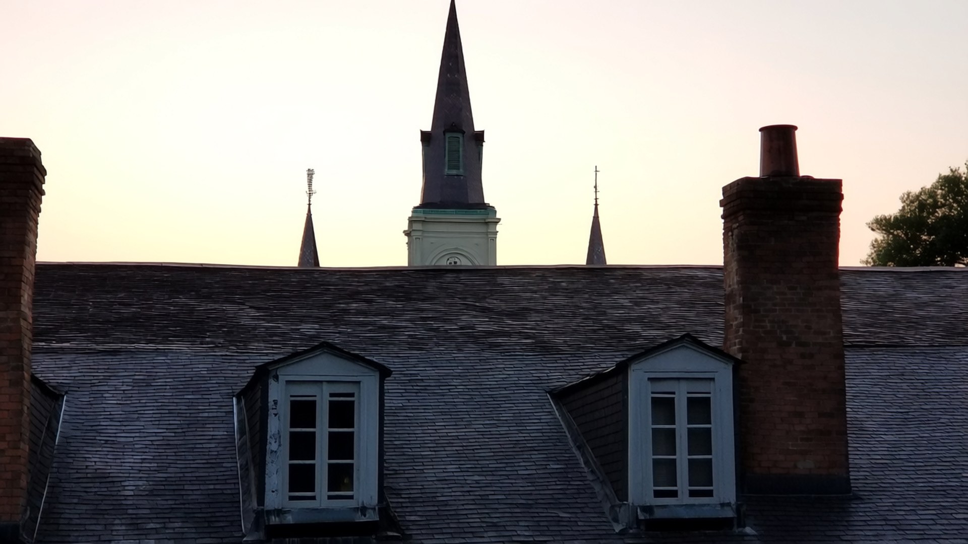 Rooftop Cathedral View 壁紙 — 帶有老虎窗和磚砌煙囪的剪影屋頂，在柔和粉彩的日落天空下襯托出教堂尖塔。