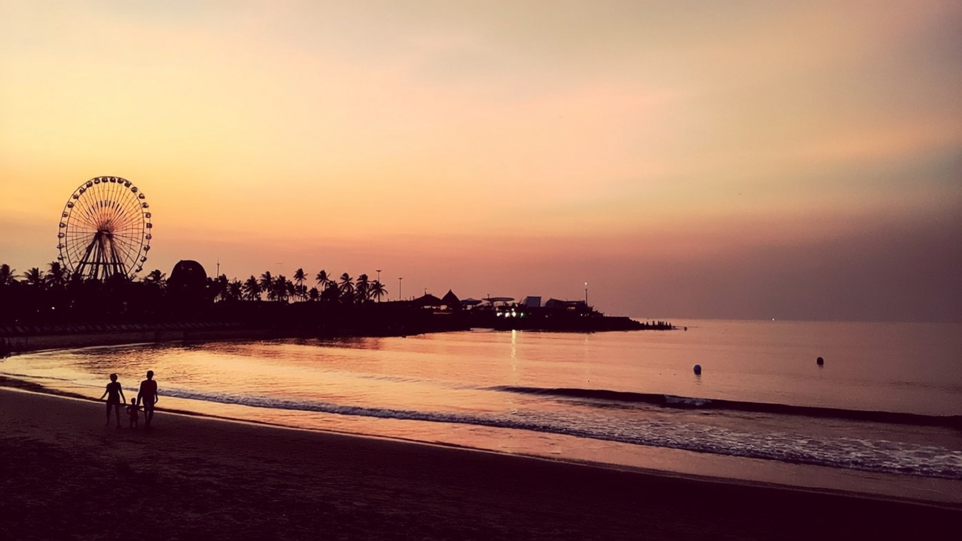 Ancol Sunset Fun 壁紙 — 日落時分，兩個人在沙灘上遛狗的剪影，遠處有摩天輪和棕櫚樹。