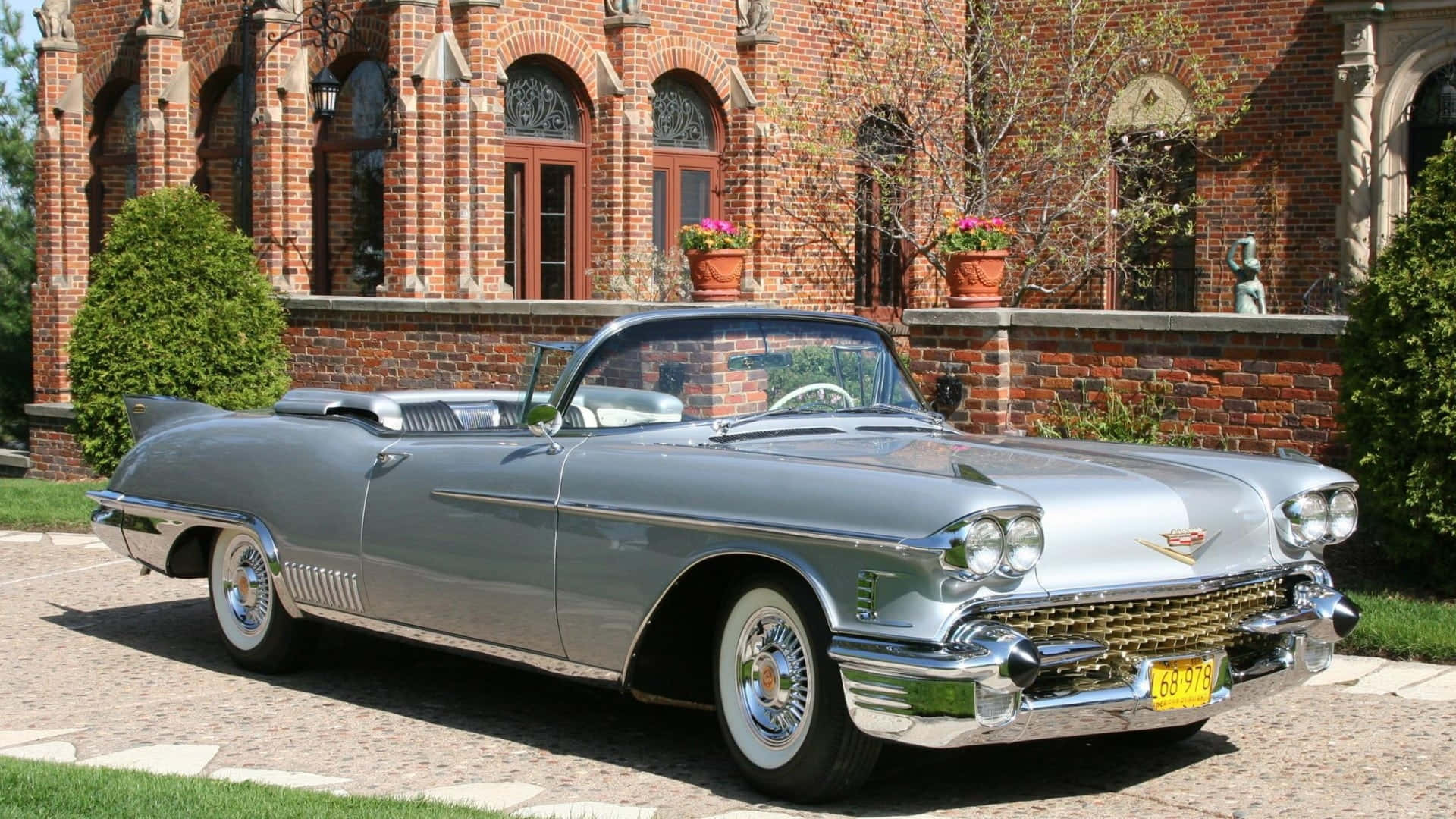 Cadillac Eldorado 桌布 壁紙 — 一輛閃亮的銀色1959年凱迪拉克敞篷車停泊在鵝卵石車道上，前面是一座宏偉的紅磚豪宅，配有修剪整齊的樹籬和盆栽花卉。