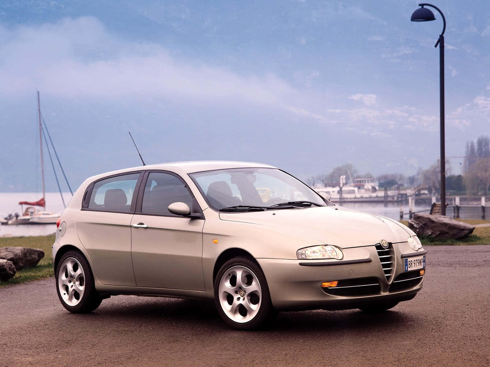 Alfa Romeo 147 桌布 壁紙 — 一輛銀色愛快羅密歐147掀背車停泊在湖邊道路上，背景有帆船和山脈，天空多雲。