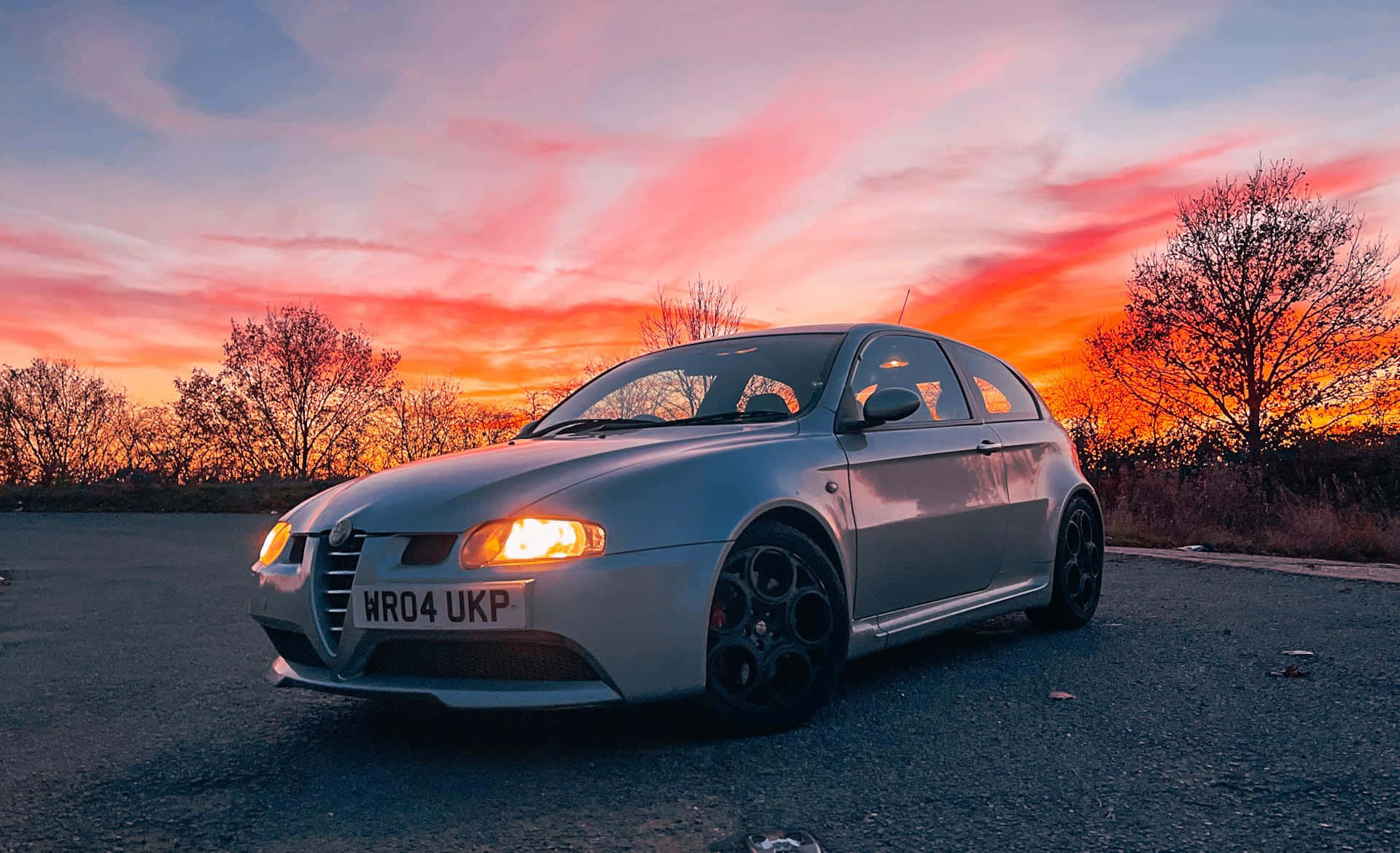 Alfa Romeo 147 桌布 壁紙 — 一輛銀色愛快羅密歐跑車停在柏油路上，背景是火紅的日落天空，襯托著光禿樹木的剪影。