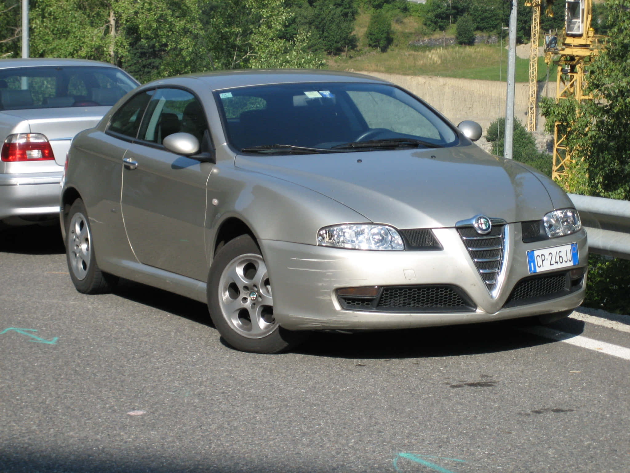 Alfa Romeo Gt 桌布 壁紙 — 一輛銀色愛快羅密歐Spider雙門跑車停放在陽光普照的路邊，背景有綠樹和黃色起重機。