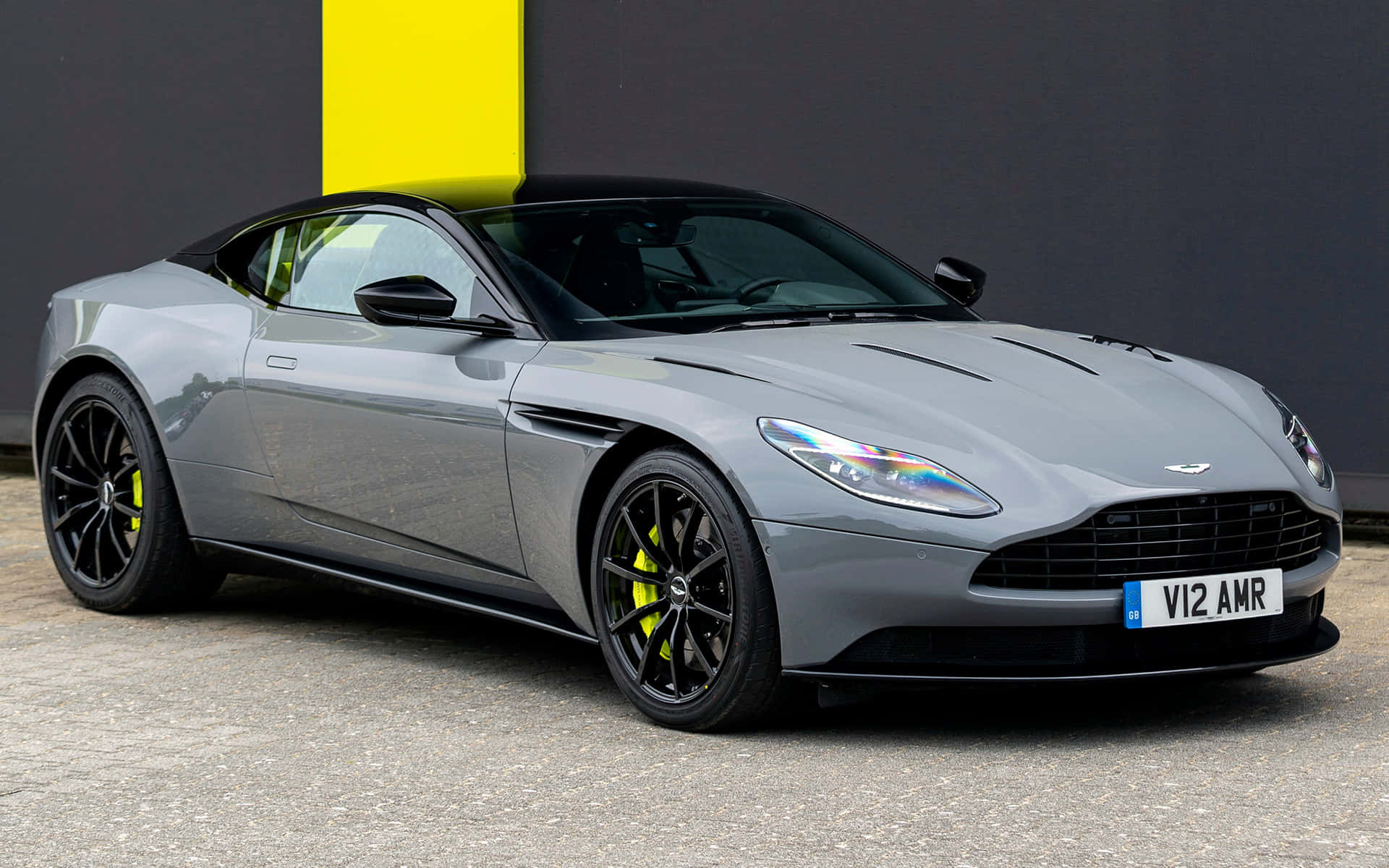 Aston Martin Db11 桌布 壁紙 — 一輛時尚的銀色 Aston Martin DB11 AMR，帶有黑色飾邊和鮮黃色煞車卡鉗，停放在大膽的黃黑牆前。