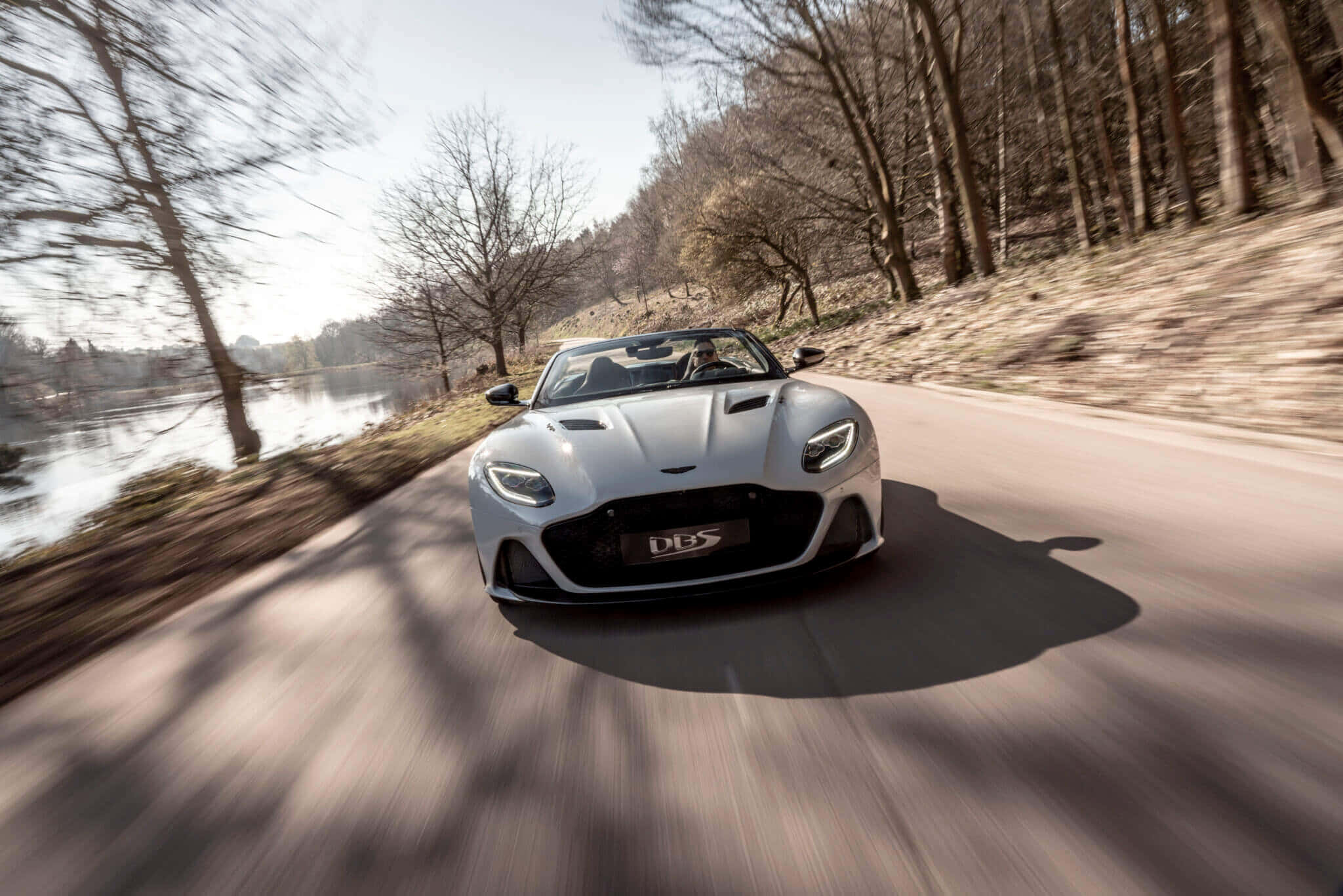 Aston Martin Dbs Superleggera 桌布 壁紙 — 一輛銀色Aston Martin DBS超級跑車沿著河邊蜿蜒的道路穿過光禿禿的冬季樹木飛馳。